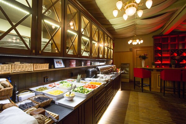 A buffet area with various food items, a counter with a coffee machine, and a bar area with red chairs.