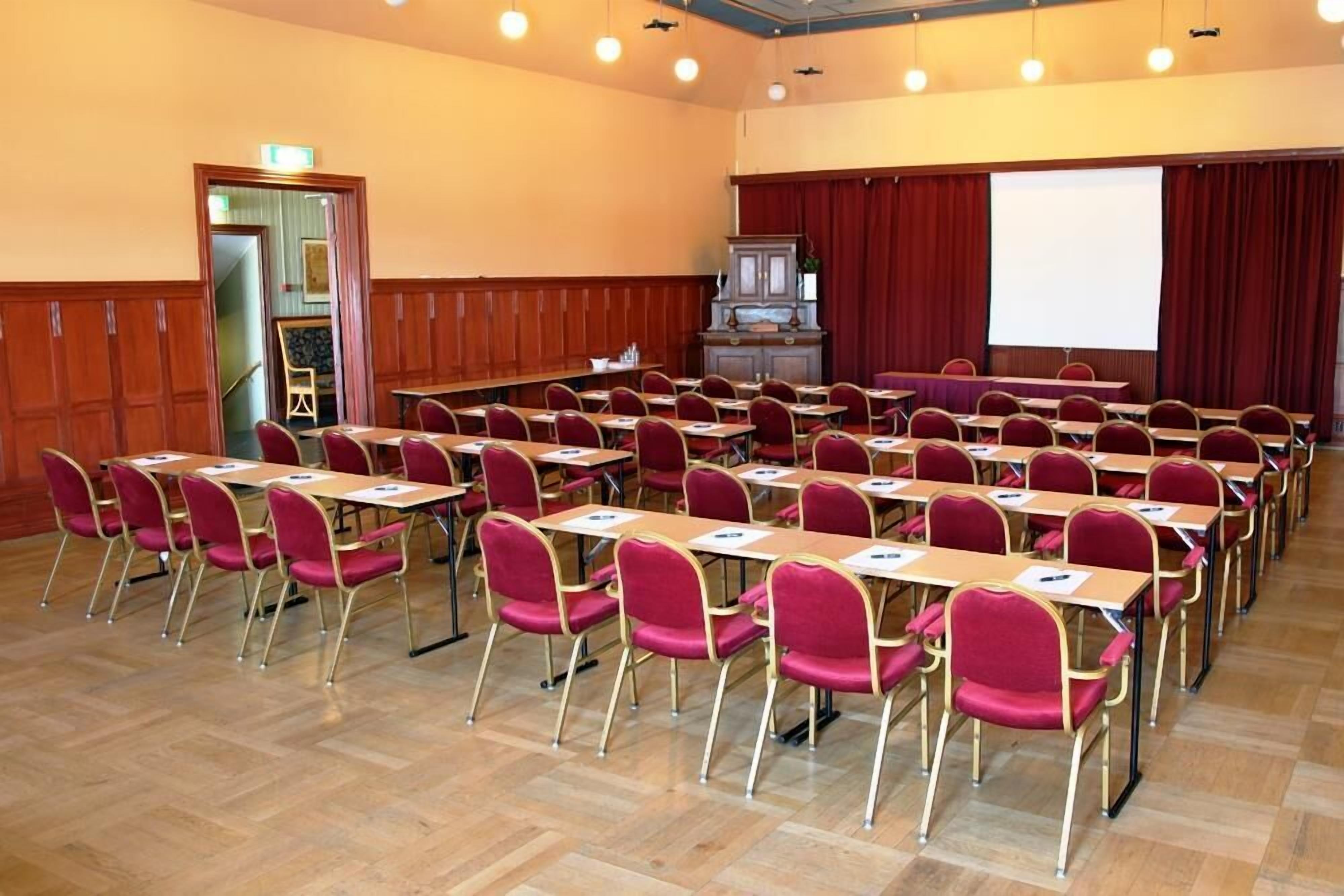 Una sala conferenze con file di tavoli e sedie, un palco con uno schermo per proiettore e una parete rivestita in legno.