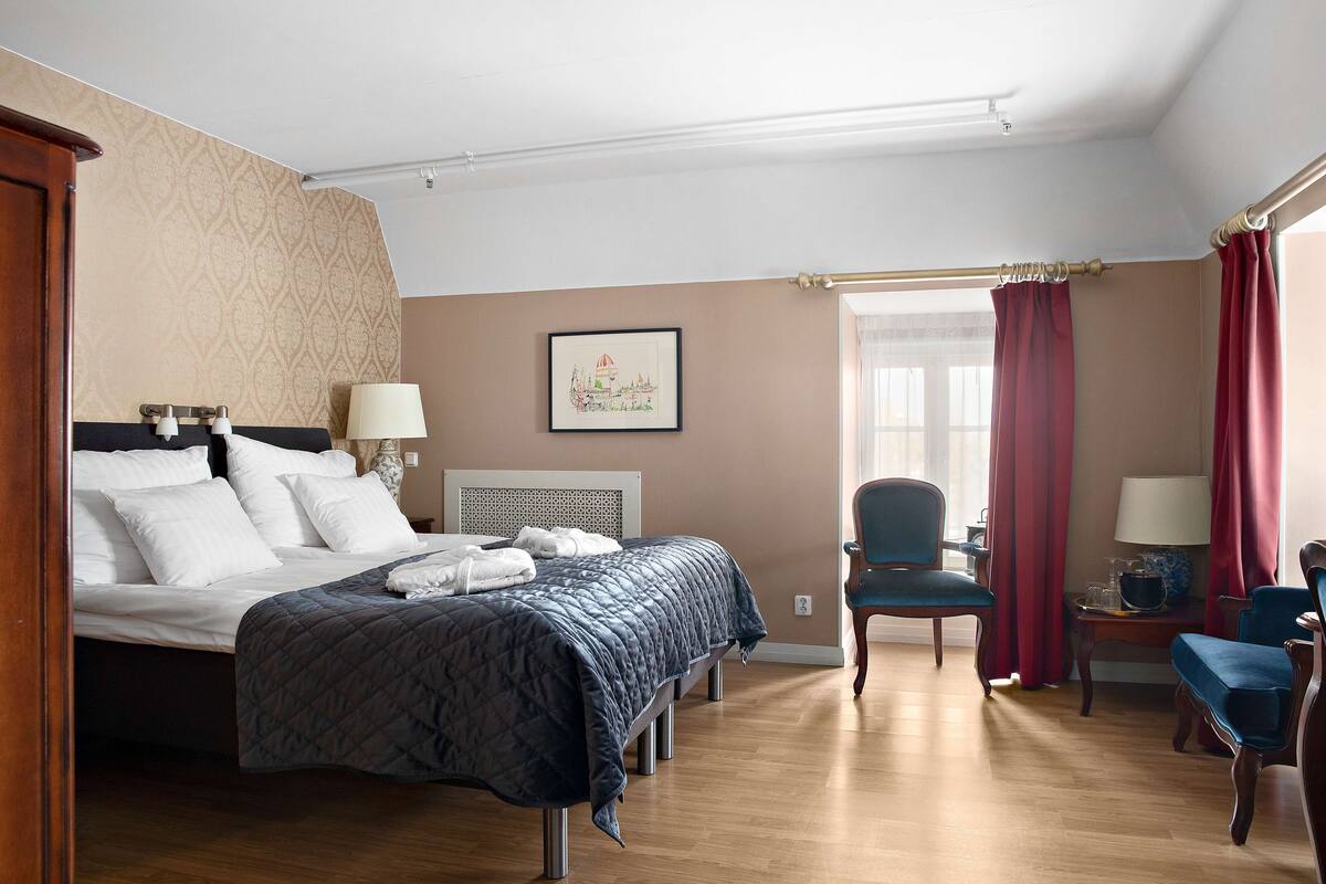 Ein Hotelzimmer mit einem großen Bett, zwei Nachttischen mit Lampen, einem Sessel und einem Fenster mit Vorhängen.