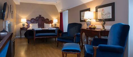 A hotel room with a bed, two blue armchairs, a wooden side table with a lamp, and a wall-mounted TV.