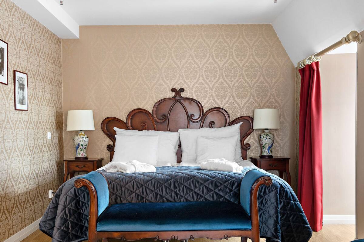 Ein Schlafzimmer mit einem hölzernen Bett, zwei Nachttischen mit Lampen, gemustertem Wandpapier und einem roten Vorhang.