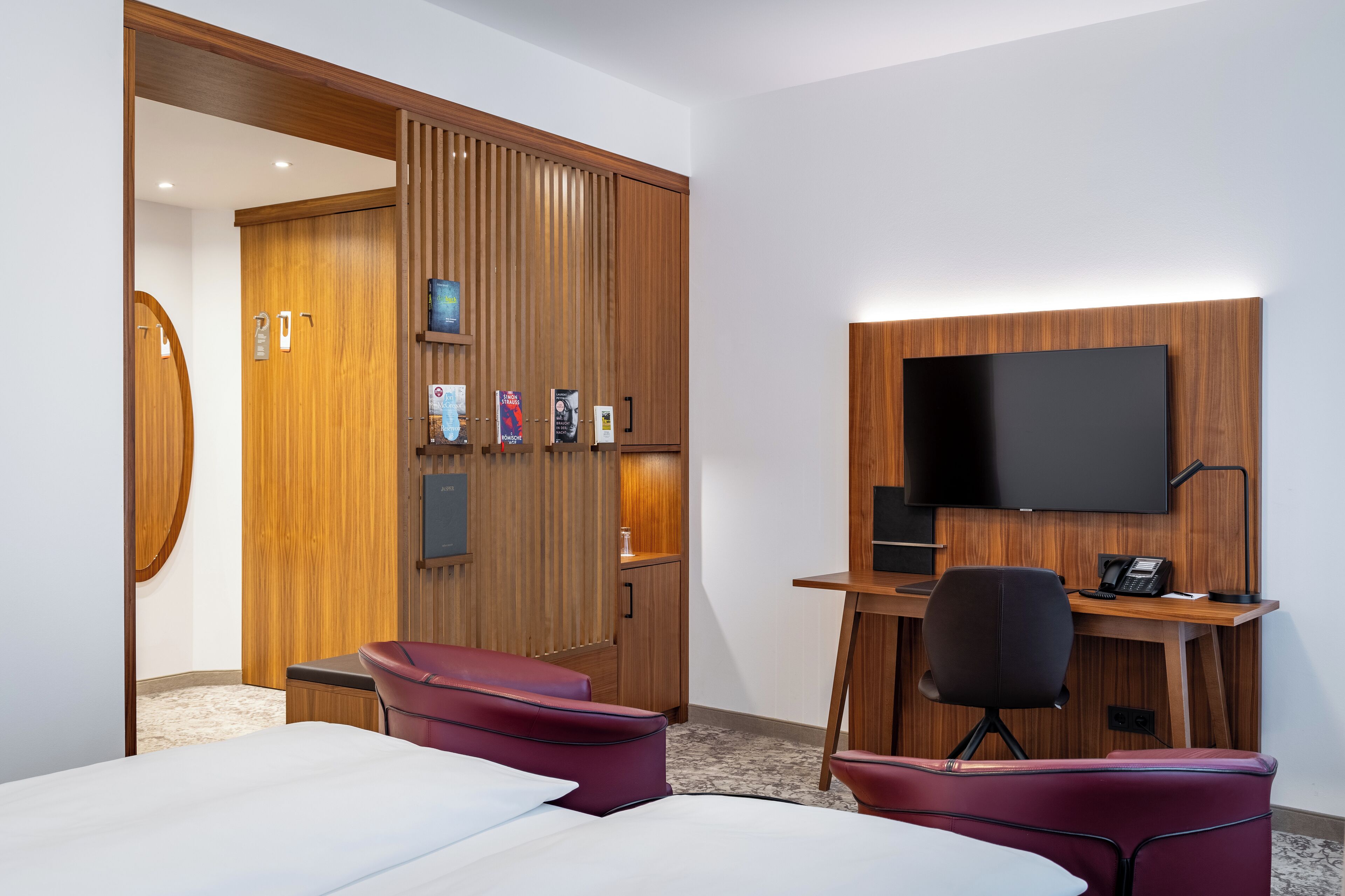 A hotel room with a wooden wardrobe, a desk with a flat-screen TV, two red chairs, and a bed.
