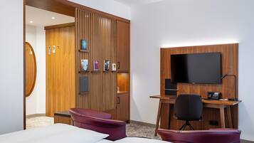 A hotel room with a wooden wardrobe, a desk with a flat-screen TV, two red chairs, and a bed.
