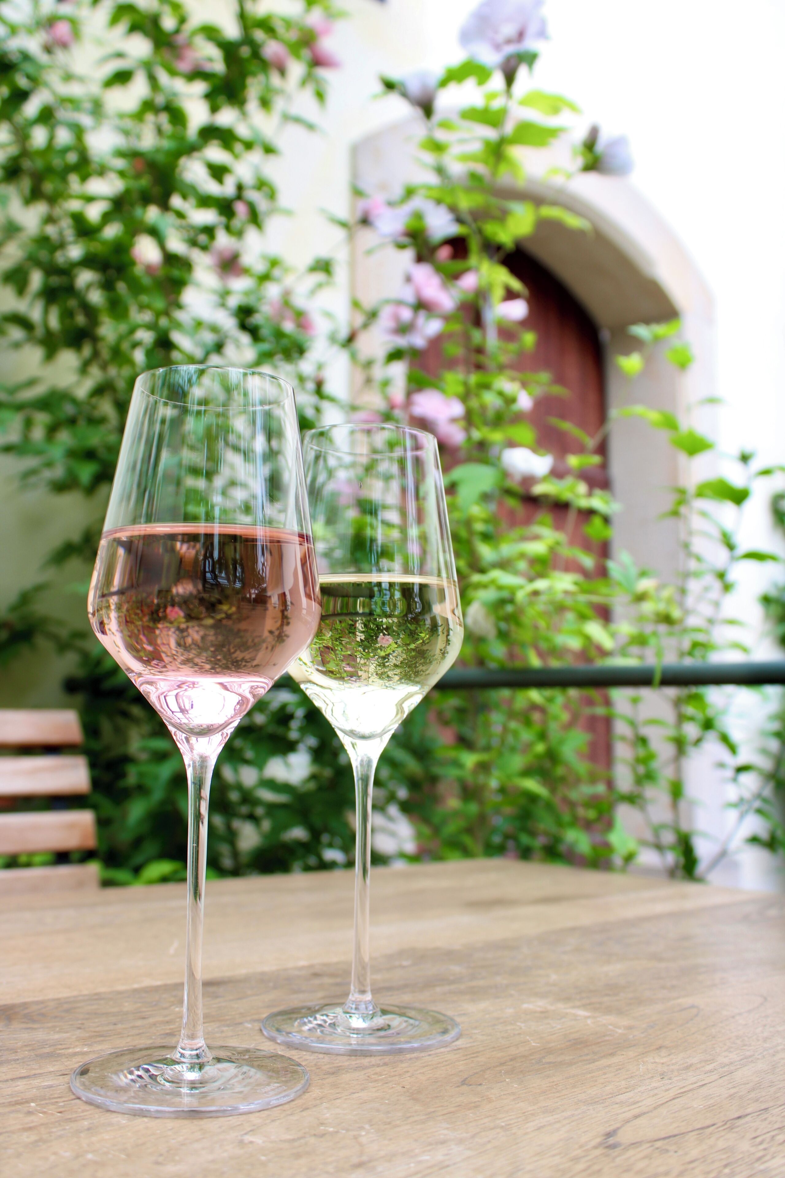 Two wine glasses on a wooden table with a view of a garden and a building in the background.