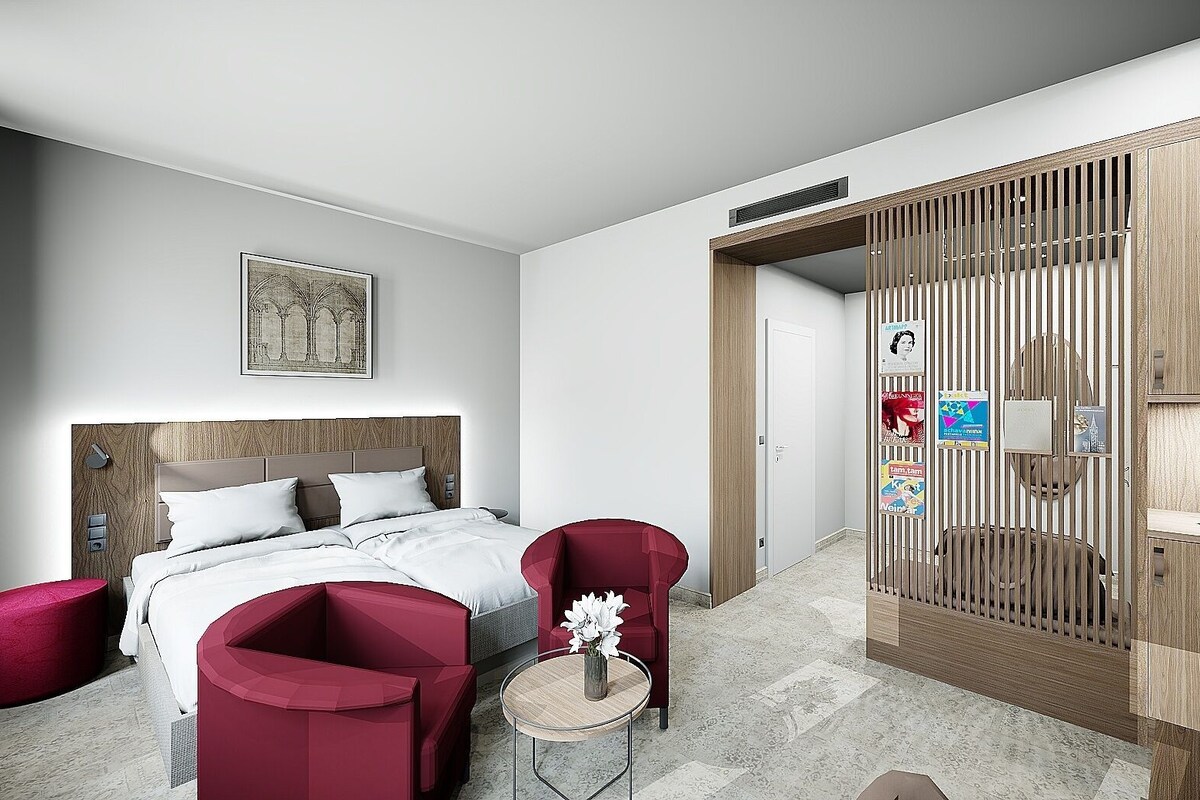 A modern hotel room with a bed, two red chairs, a small round table with a vase, a wooden wardrobe, and a framed picture on the wall.