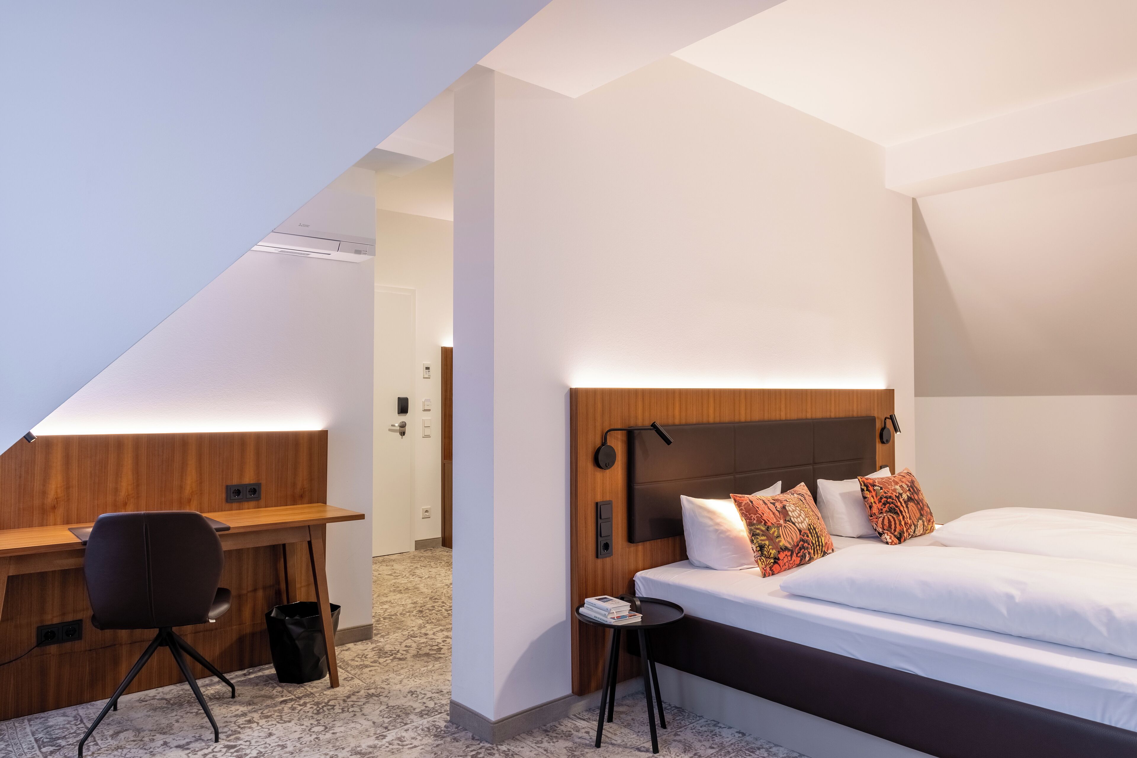 A modern hotel room with a wooden desk, a bed with white bedding, and a small side table.
