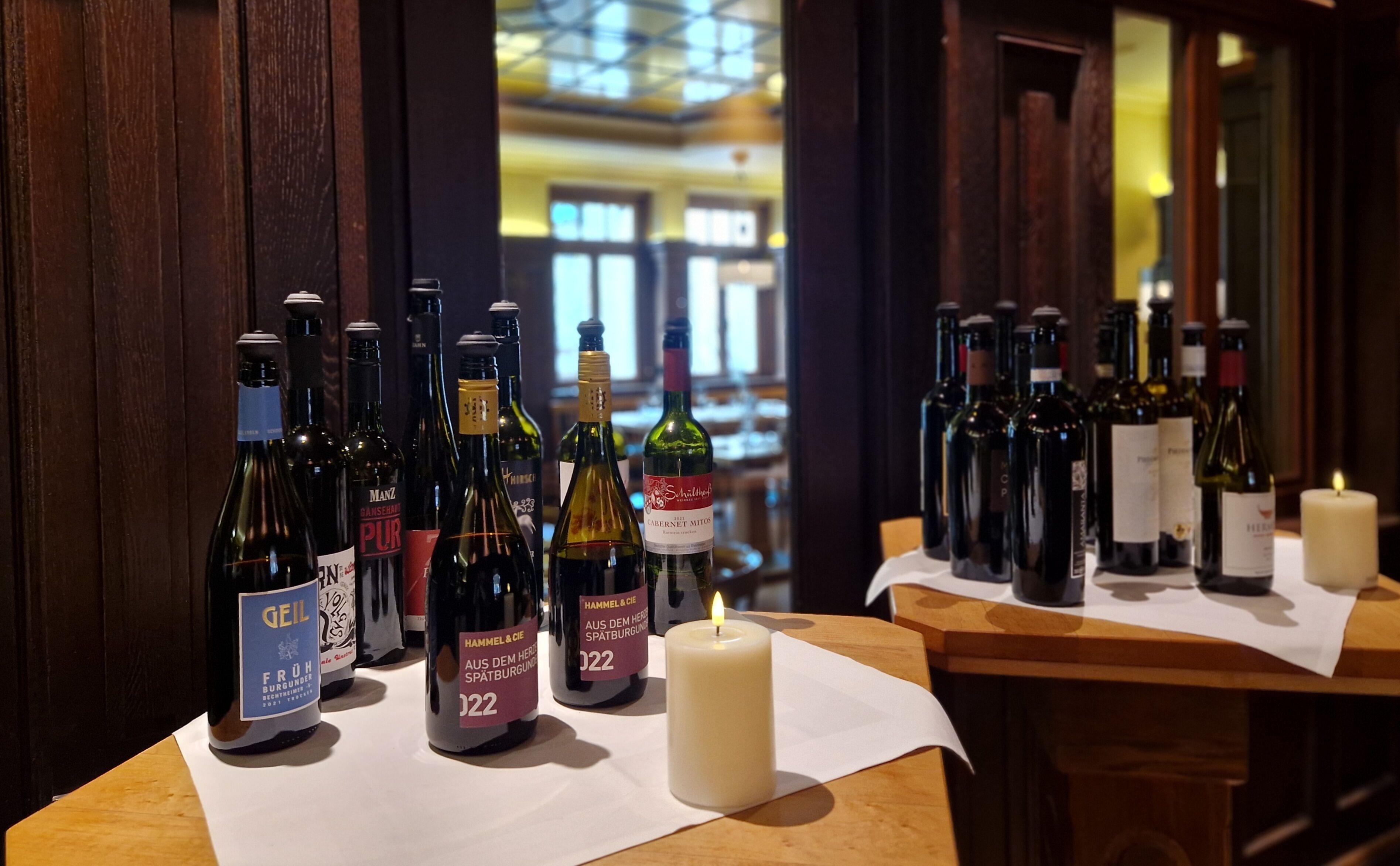 Varias botellas de vino sobre una mesa de madera, con una vela encendida, en una habitación tenuemente iluminada con paredes de madera y un vistazo a un interior luminoso al fondo.