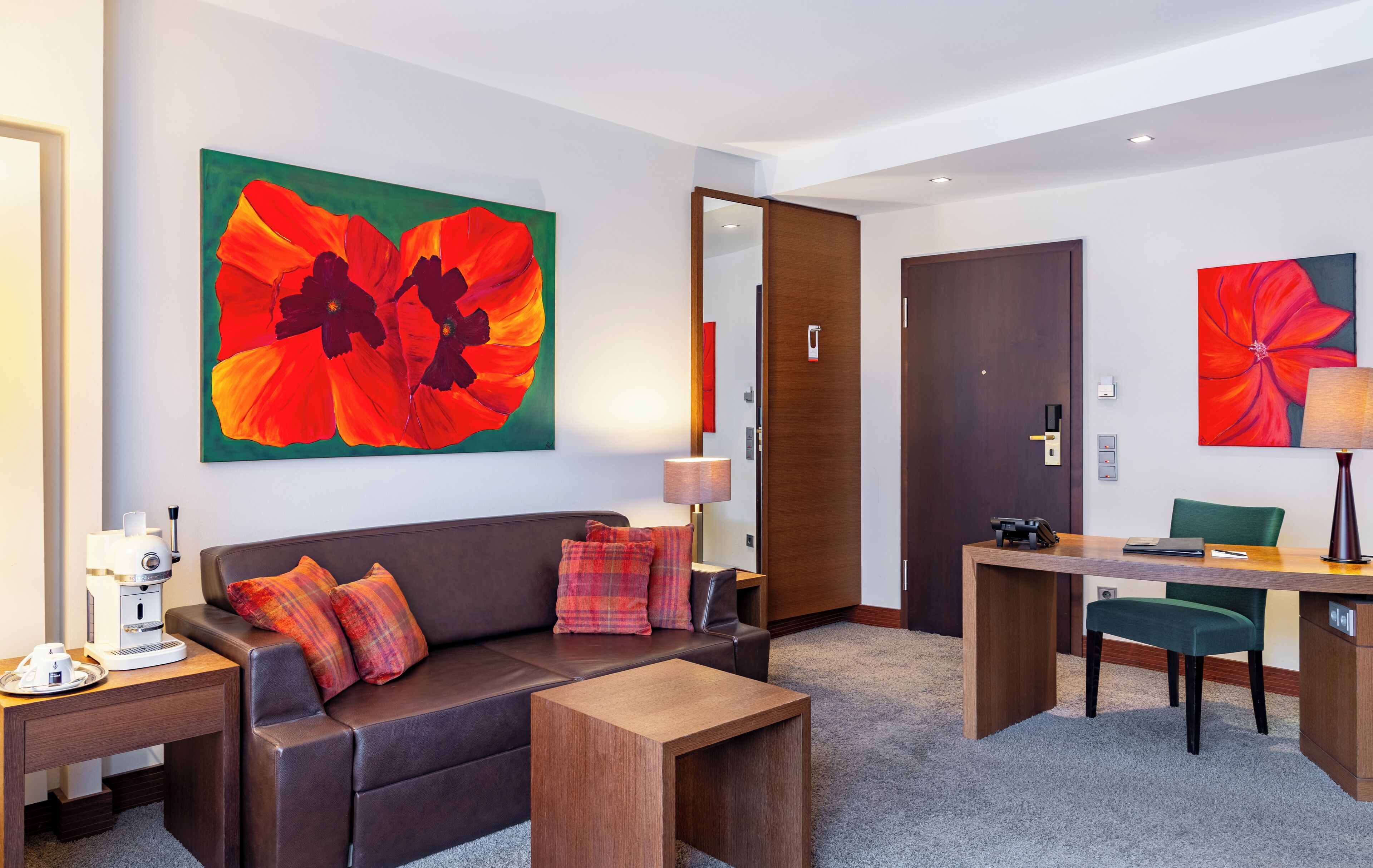 A modern hotel room with a sofa, coffee table, desk, and chair. There are two large floral paintings on the walls.
