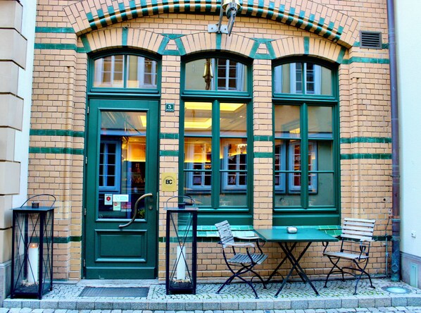A building with green-framed windows and doors, a brick facade, and outdoor seating with tables and chairs.