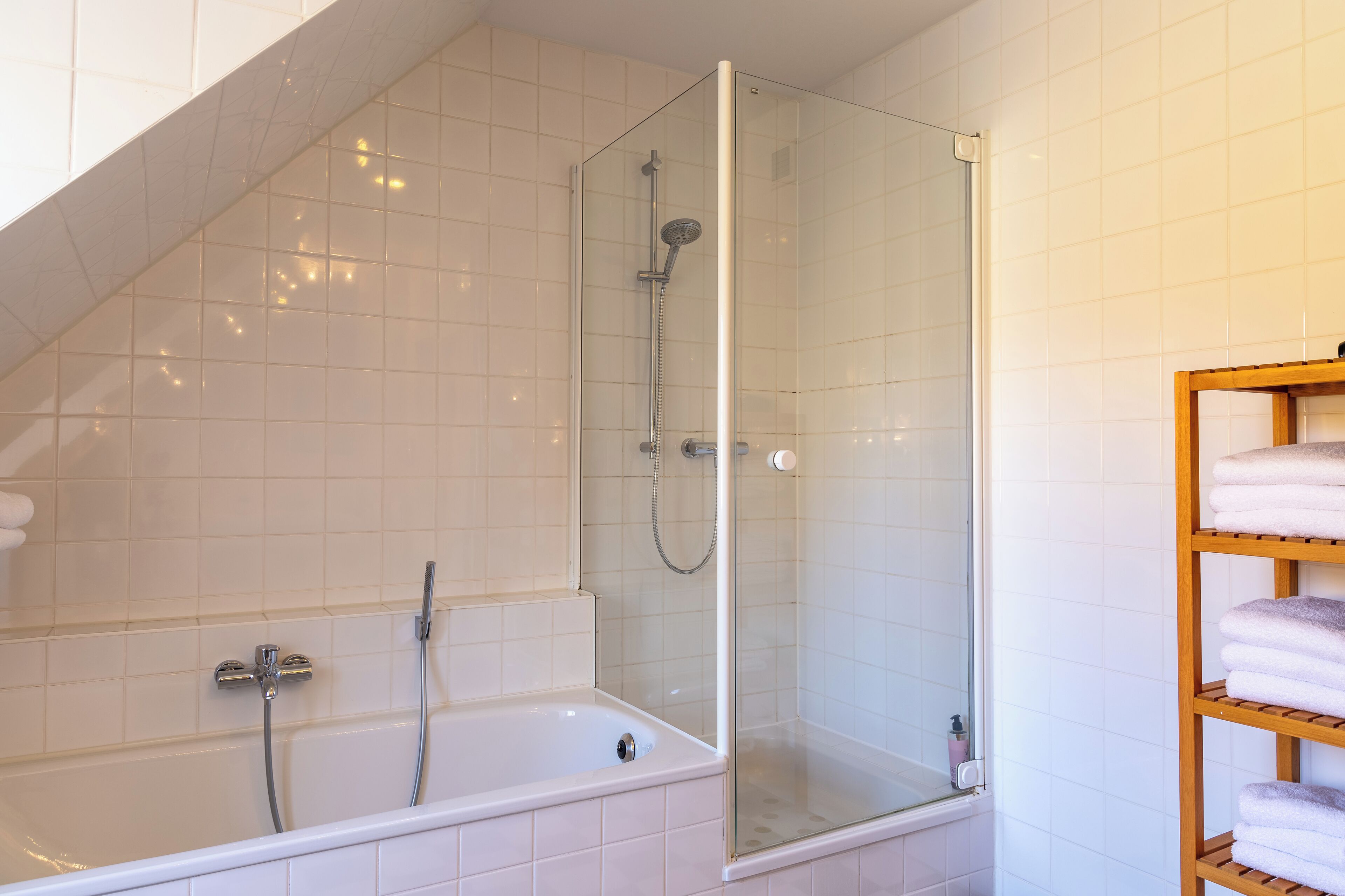 A bathroom with a glass-enclosed shower, a white bathtub, and a shelf with folded towels.