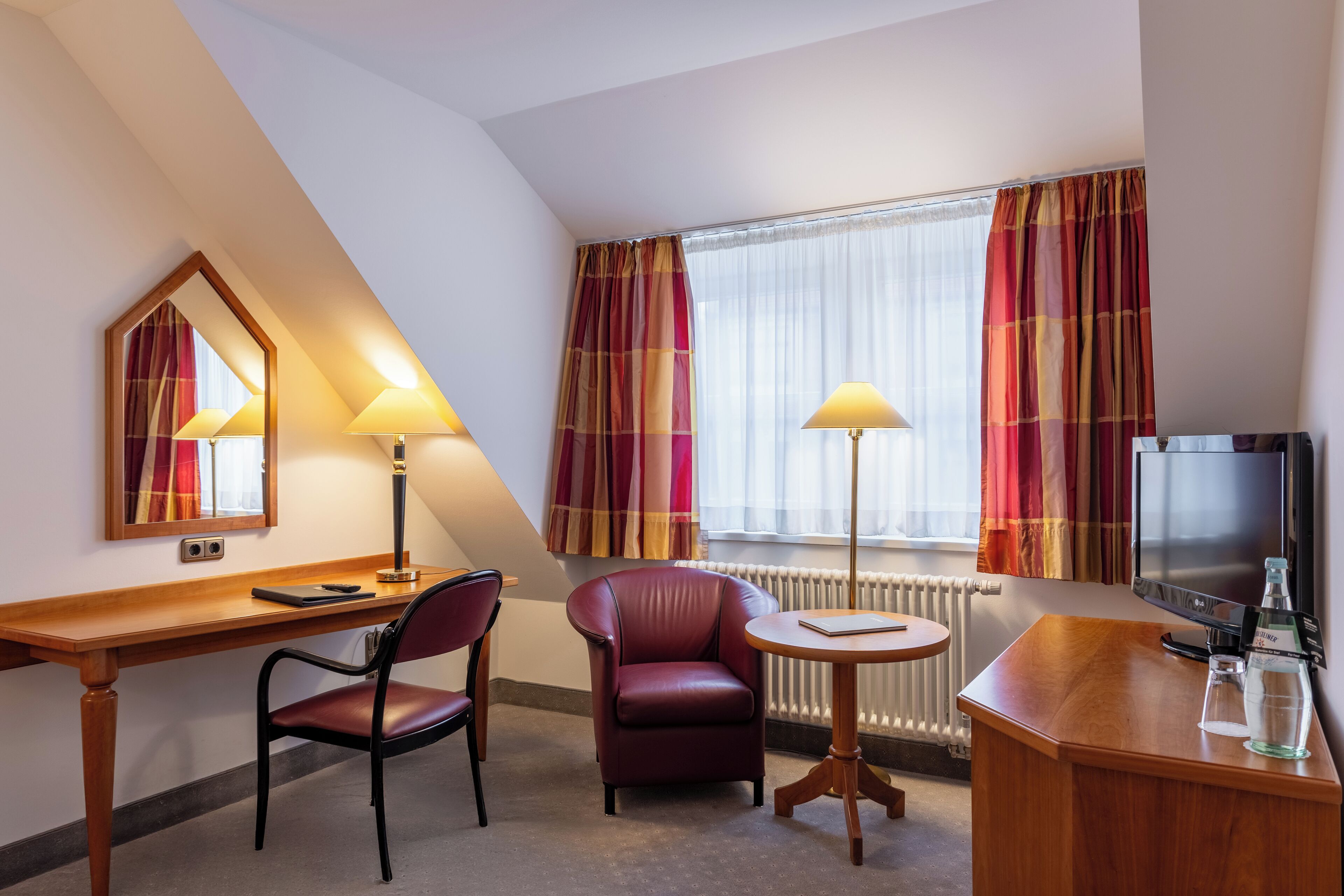 Habitación de hotel con un escritorio de madera, un sillón de cuero rojo, una mesita redonda pequeña, un televisor y un radiador.