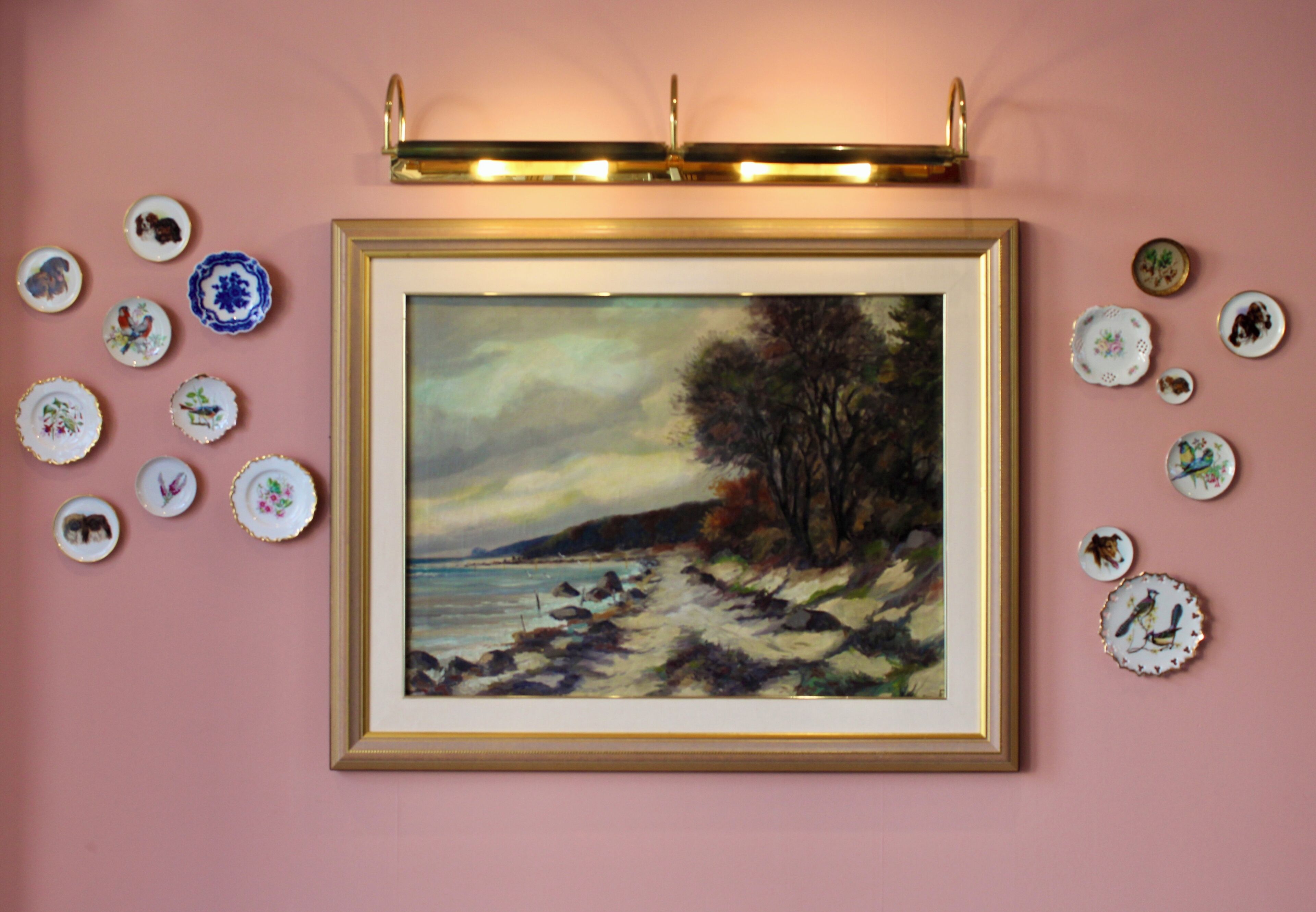Ein gerahmtes Gemälde einer Strandansicht an einer rosa Wand, umgeben von dekorativen Tellern.