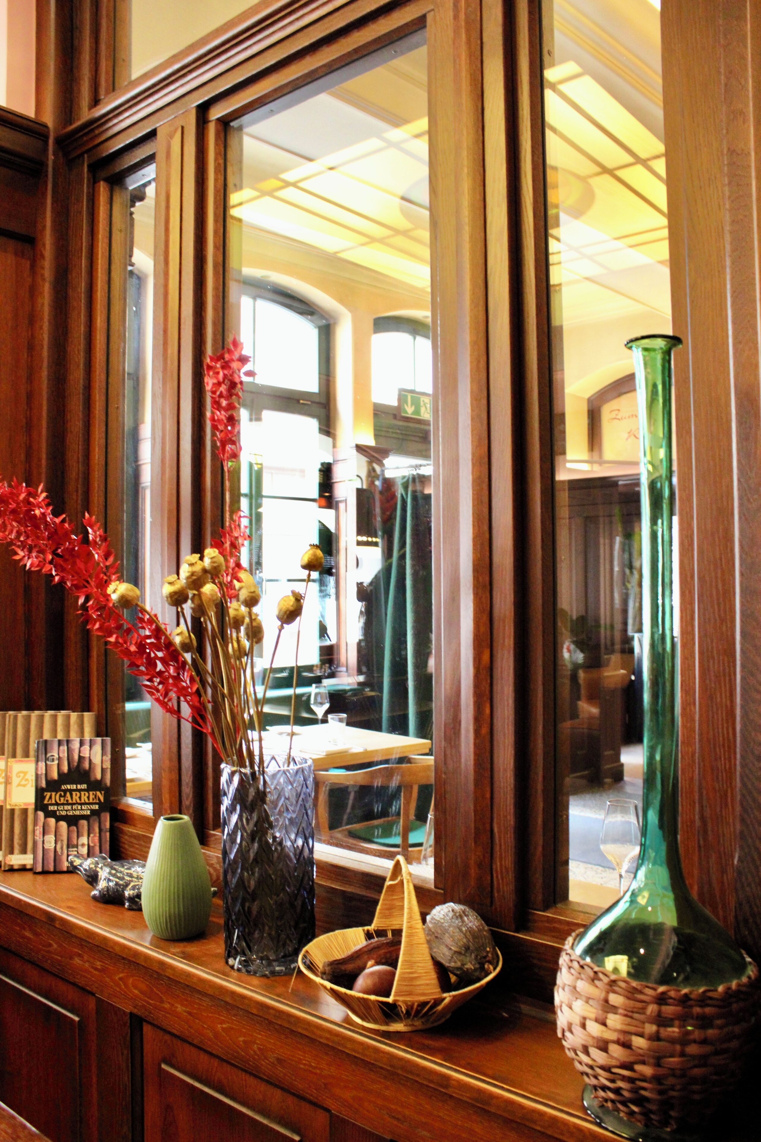 A wooden counter with a mirror reflecting a room. On the counter, there are various items including a green vase, a wicker basket, and a small wooden bowl with a coconut.