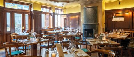 A restaurant with wooden tables and chairs, a fireplace, and large windows.