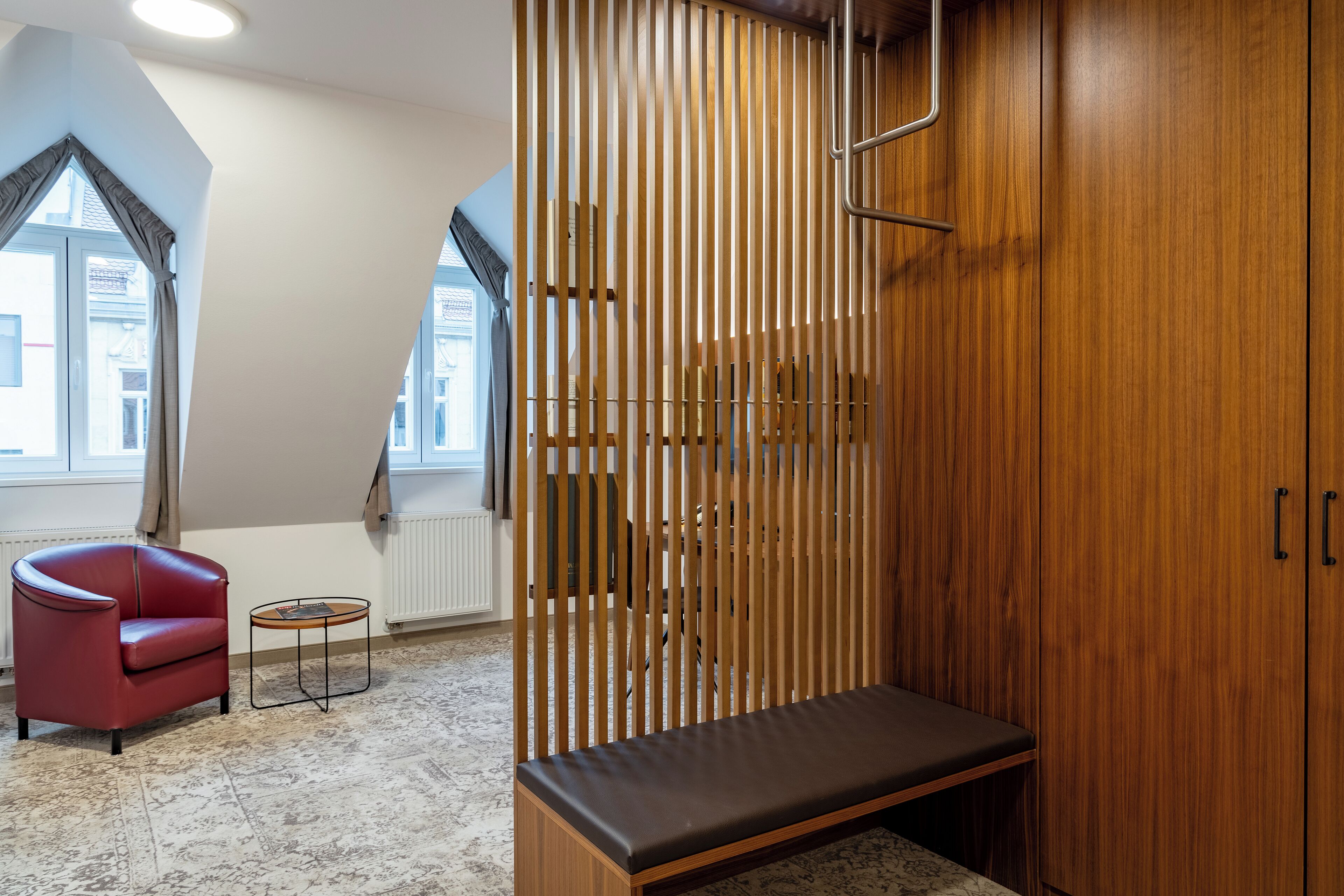 Una habitación con un armario de madera, una silla de cuero rojo, una mesita redonda y un banco.