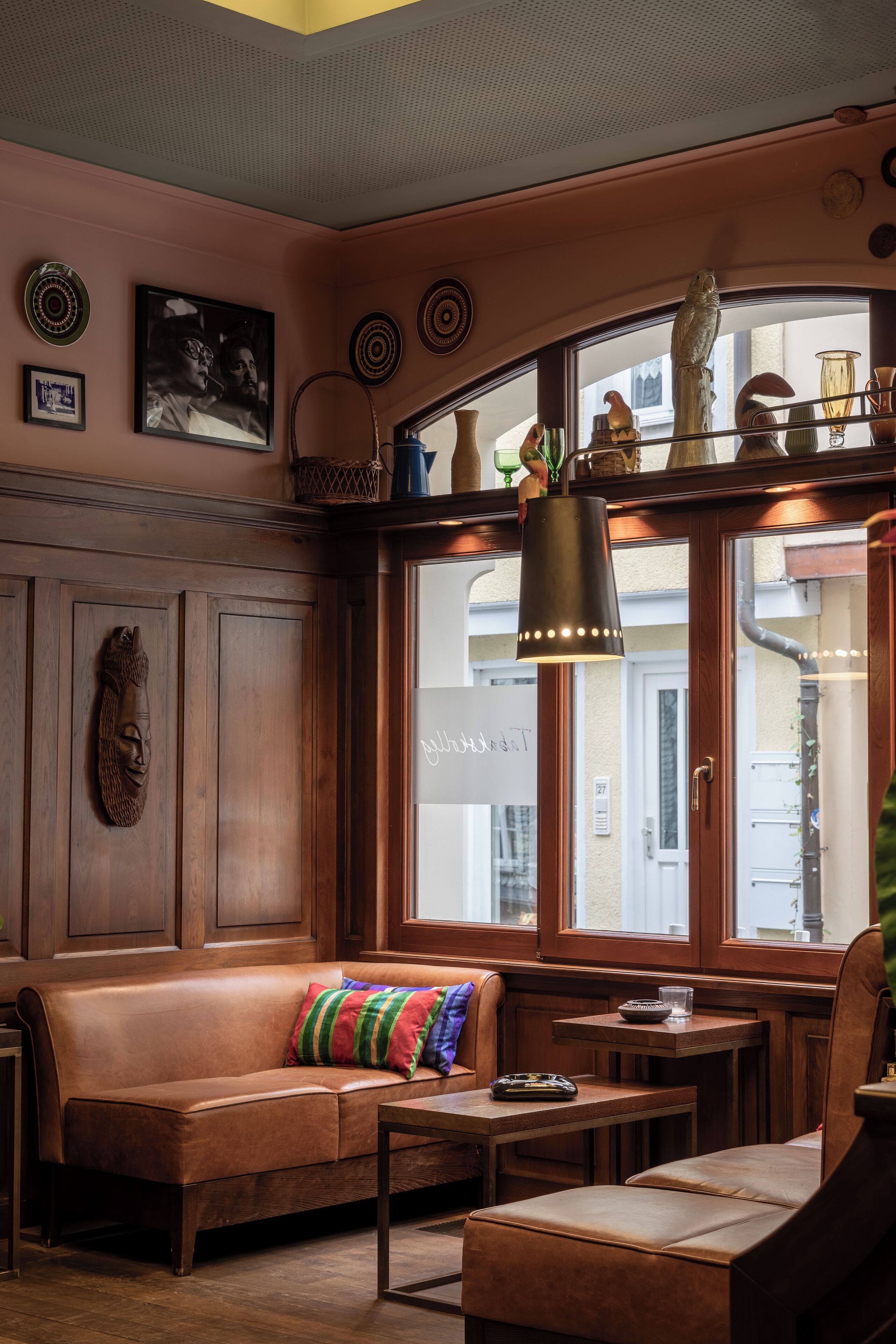 Un intérieur chaleureux avec un canapé en cuir, une petite table et un banc. Des objets décoratifs sont disposés sur des étagères, et une grande fenêtre offre une vue sur la rue.