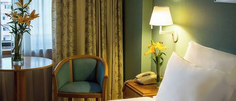 A hotel room with a bed, a chair, a bedside table with a lamp, a telephone, and a vase with flowers.