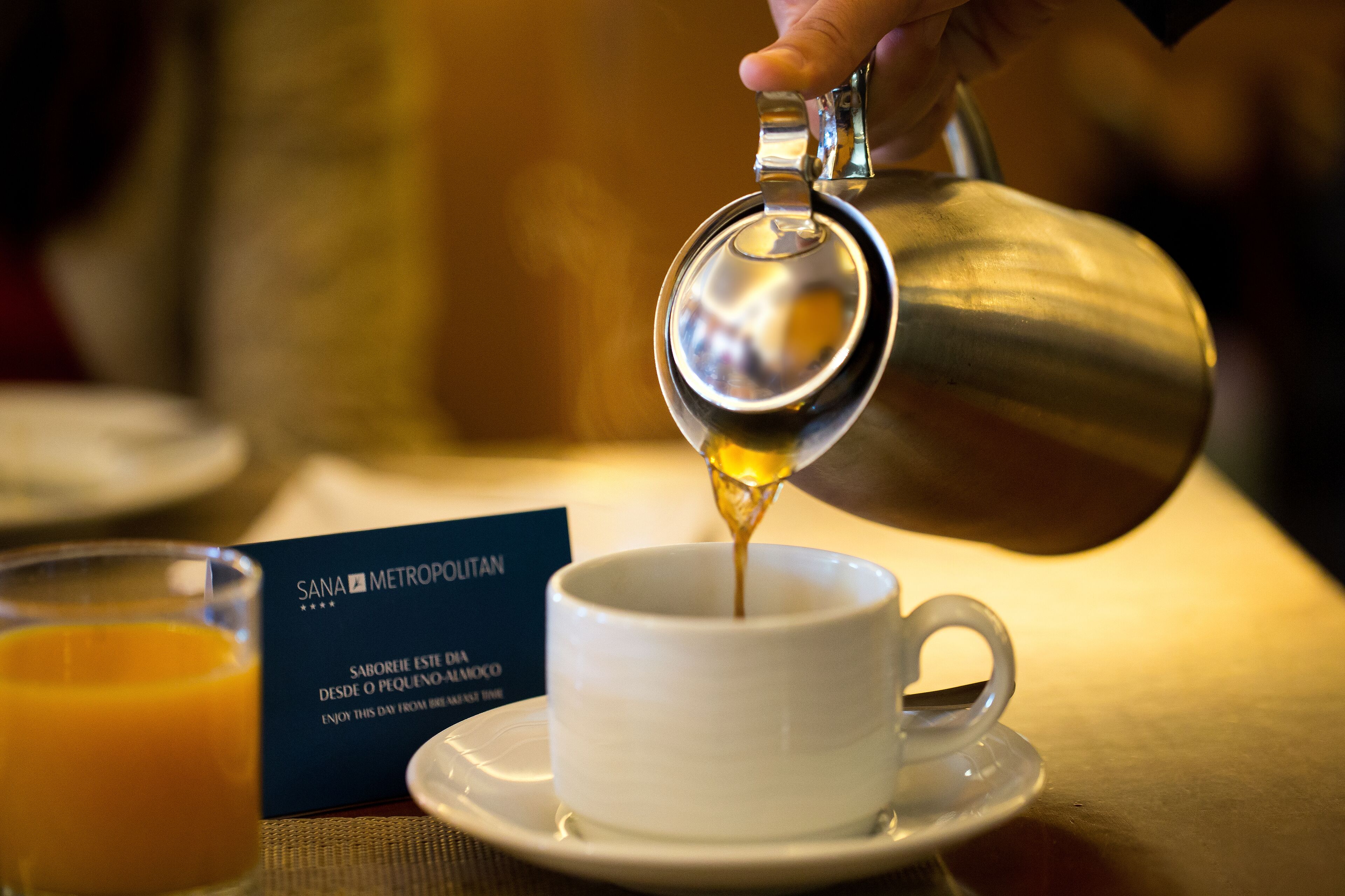 A hand pouring a golden liquid into a white cup on a saucer. A glass of orange juice and a sign with text are visible in the background.