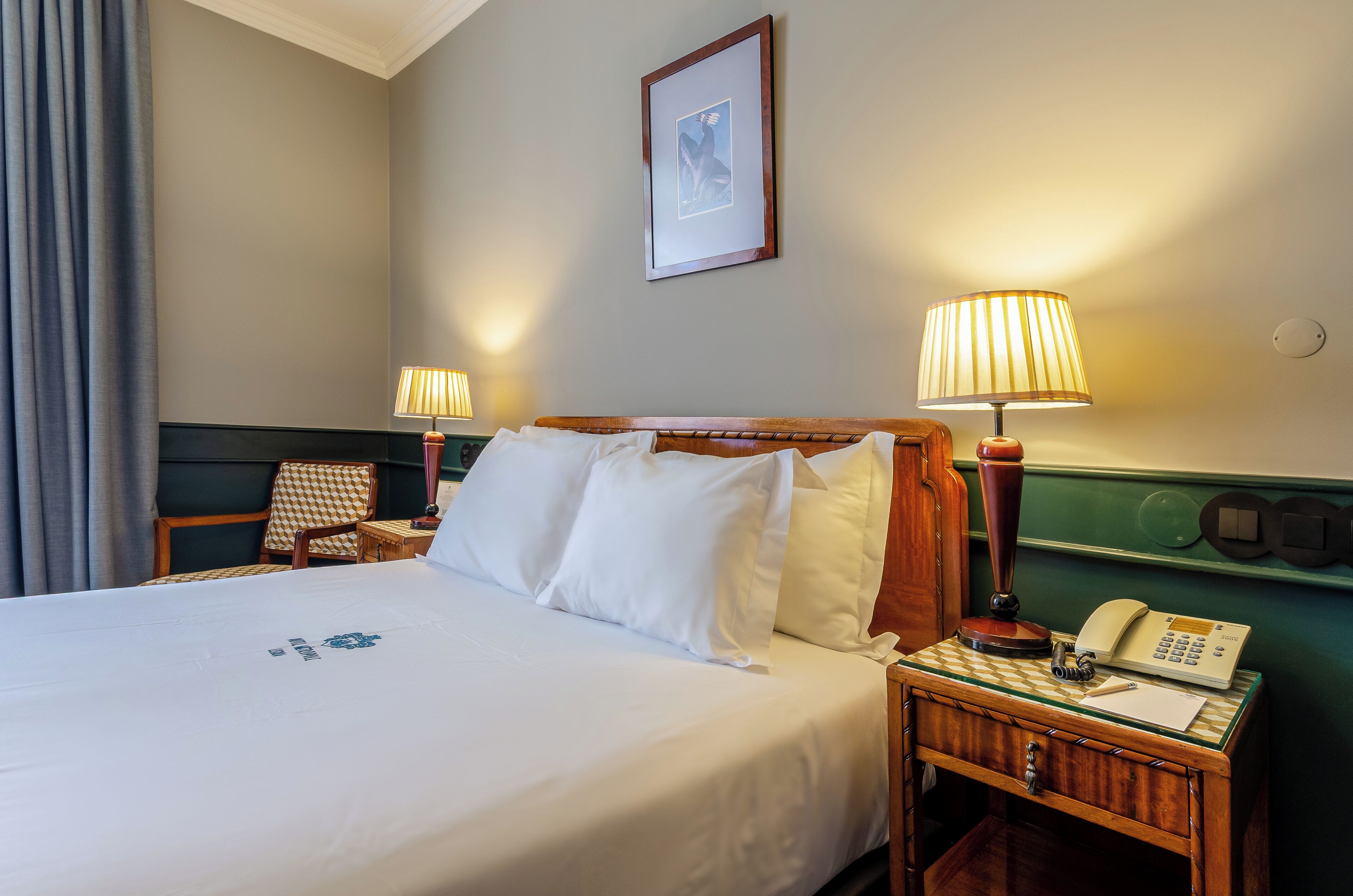 Ein Hotelzimmer mit einem ordentlich bezogenen Bett, zwei Nachttischlampen, einem Telefon, einem Sessel und einem gerahmten Bild an der Wand.