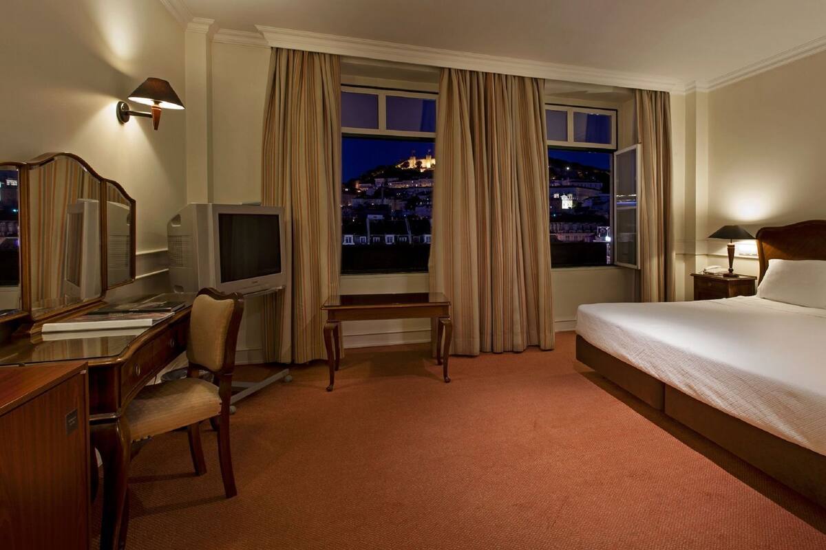 A hotel room with a bed, desk, chair, television, and a view of a cityscape at night.
