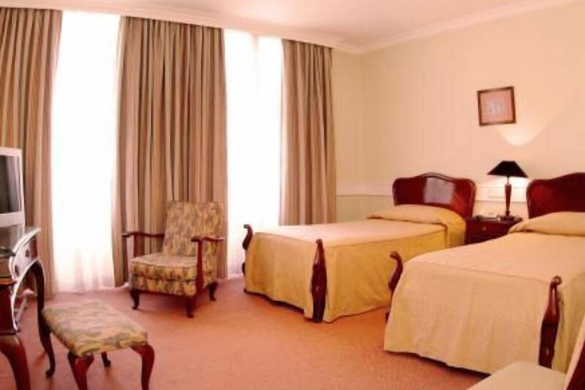 A hotel room with two beds, a chair, a bench, a TV, and curtains.