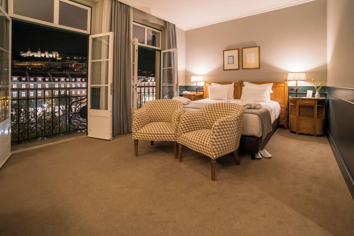 A hotel room with a large window, a bed, two checkered armchairs, a nightstand, and a lamp.