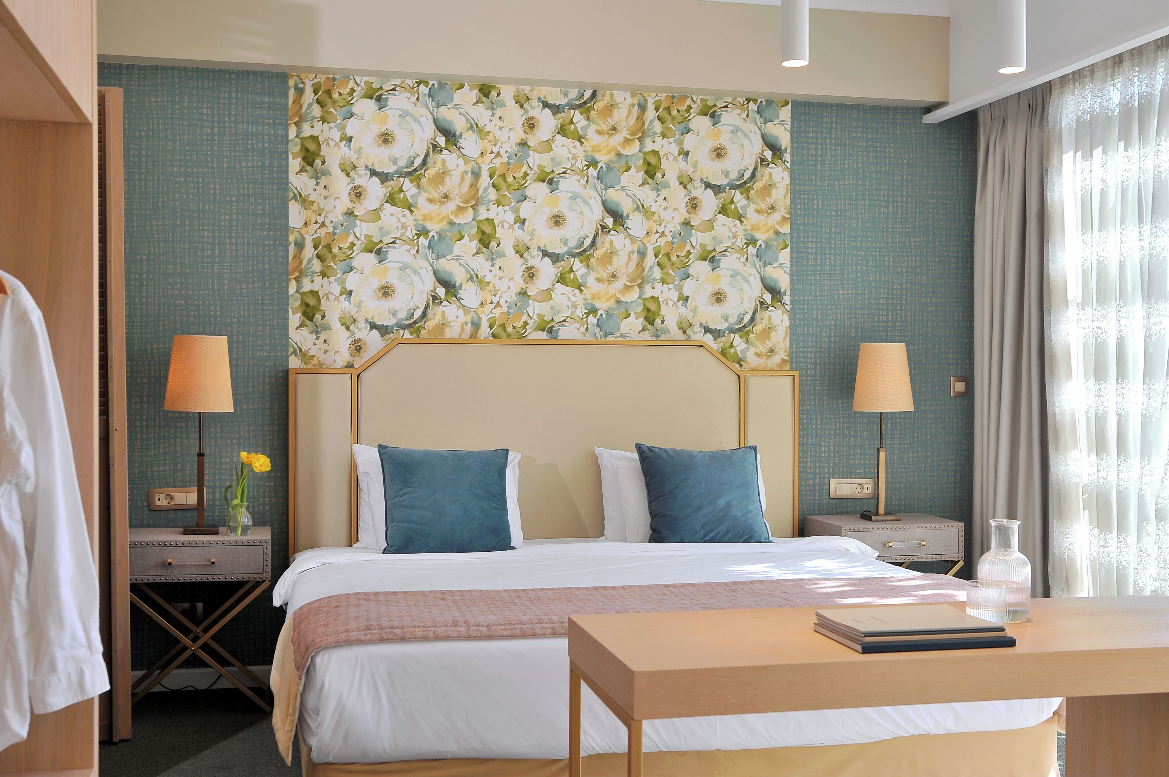 Una camera d'albergo con un letto, due comodini, una scrivania, una lampada, un vaso con fiori e un ampio carta da parati floreale.