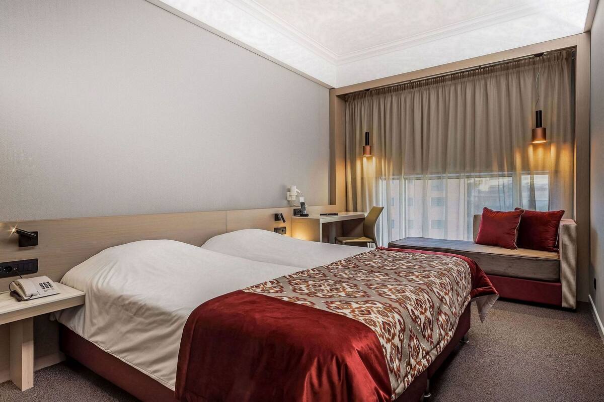 A hotel room with a bed, bedside table, lamp, and a window with curtains.