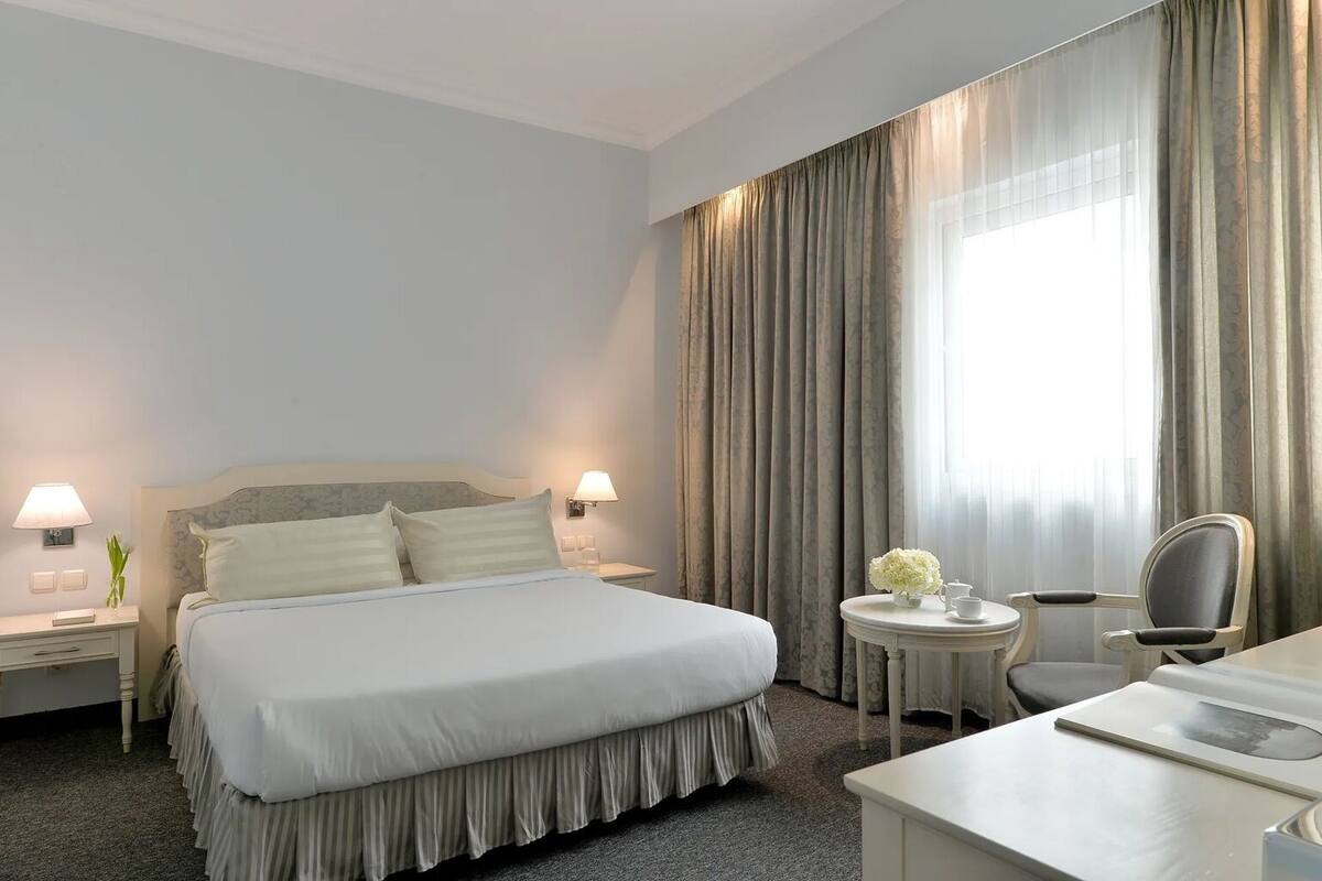 A hotel room with a large bed, two bedside tables with lamps, a round side table with a vase, a chair, and a desk with a chair.