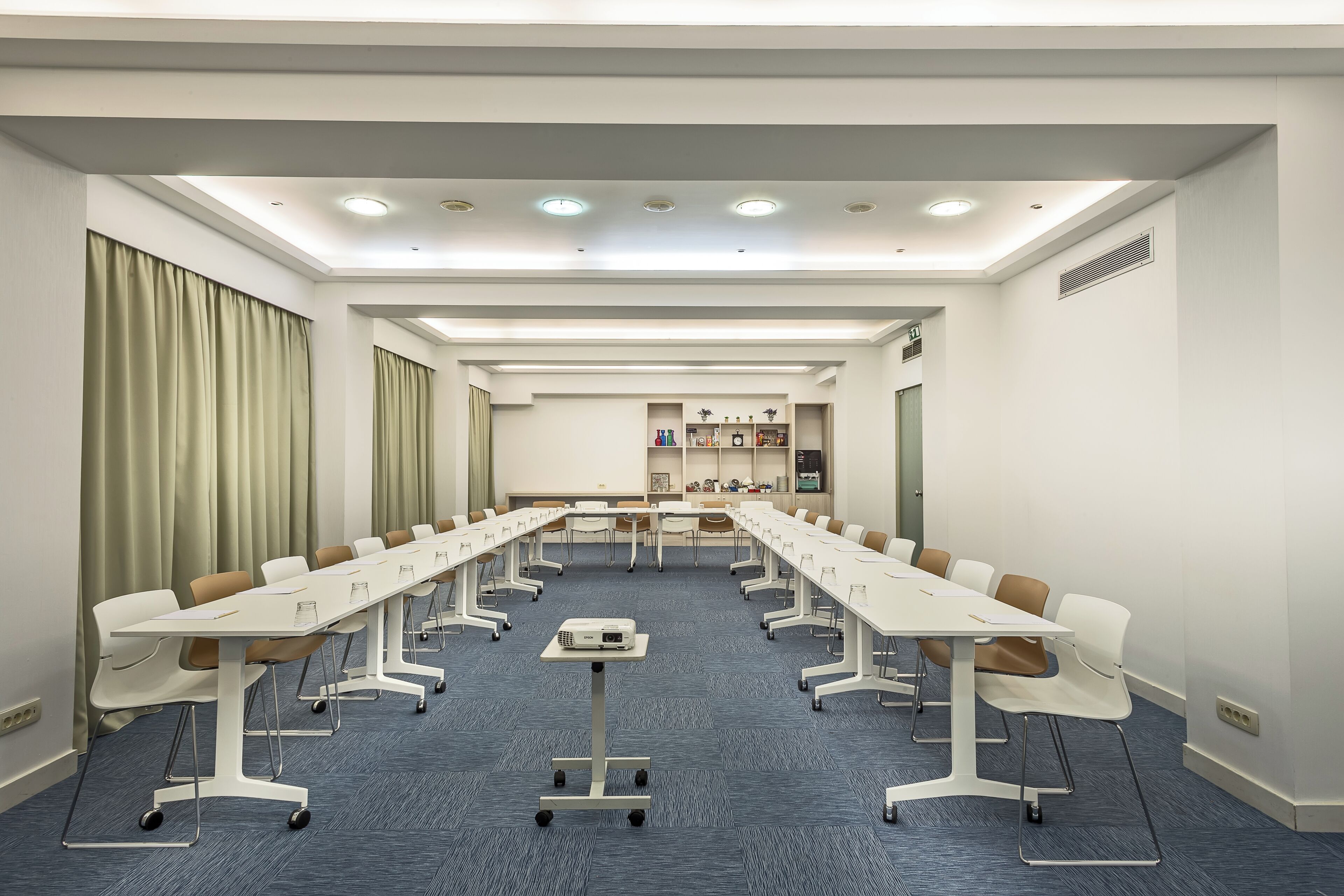 Een vergaderzaal met lange tafels, stoelen, een projectiescherm en een tapijtvloer.