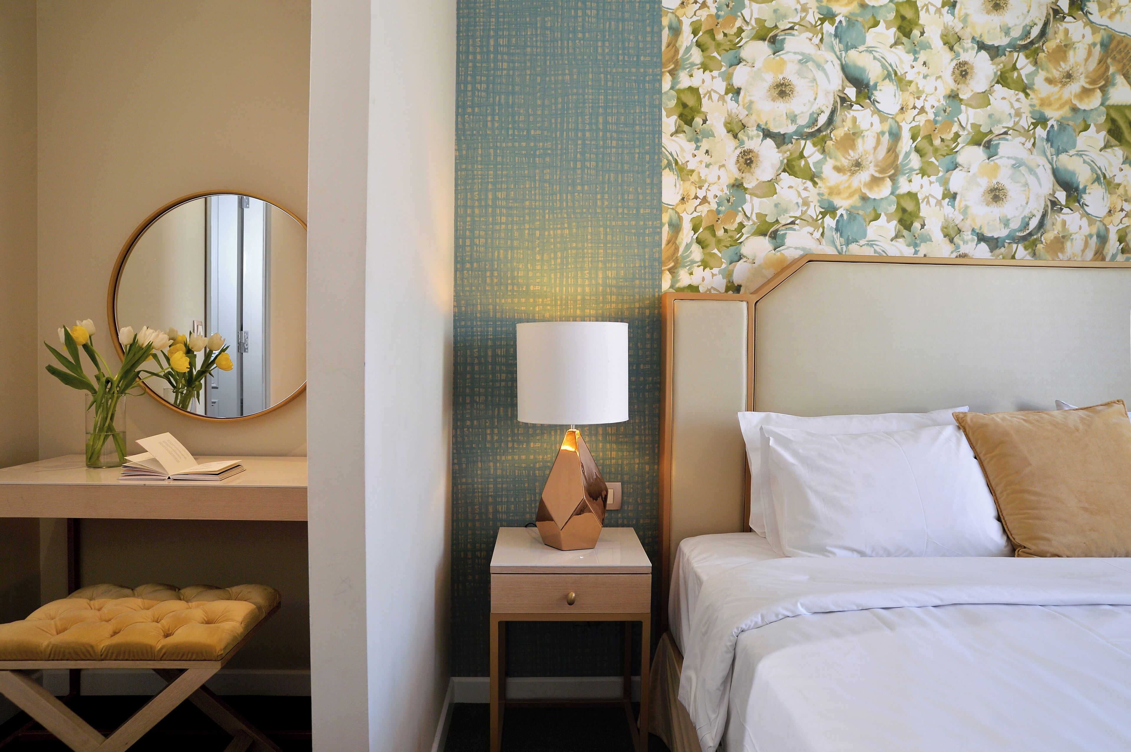 Una habitación de hotel con cama, mesita de noche, lámpara, espejo y un pequeño taburete.