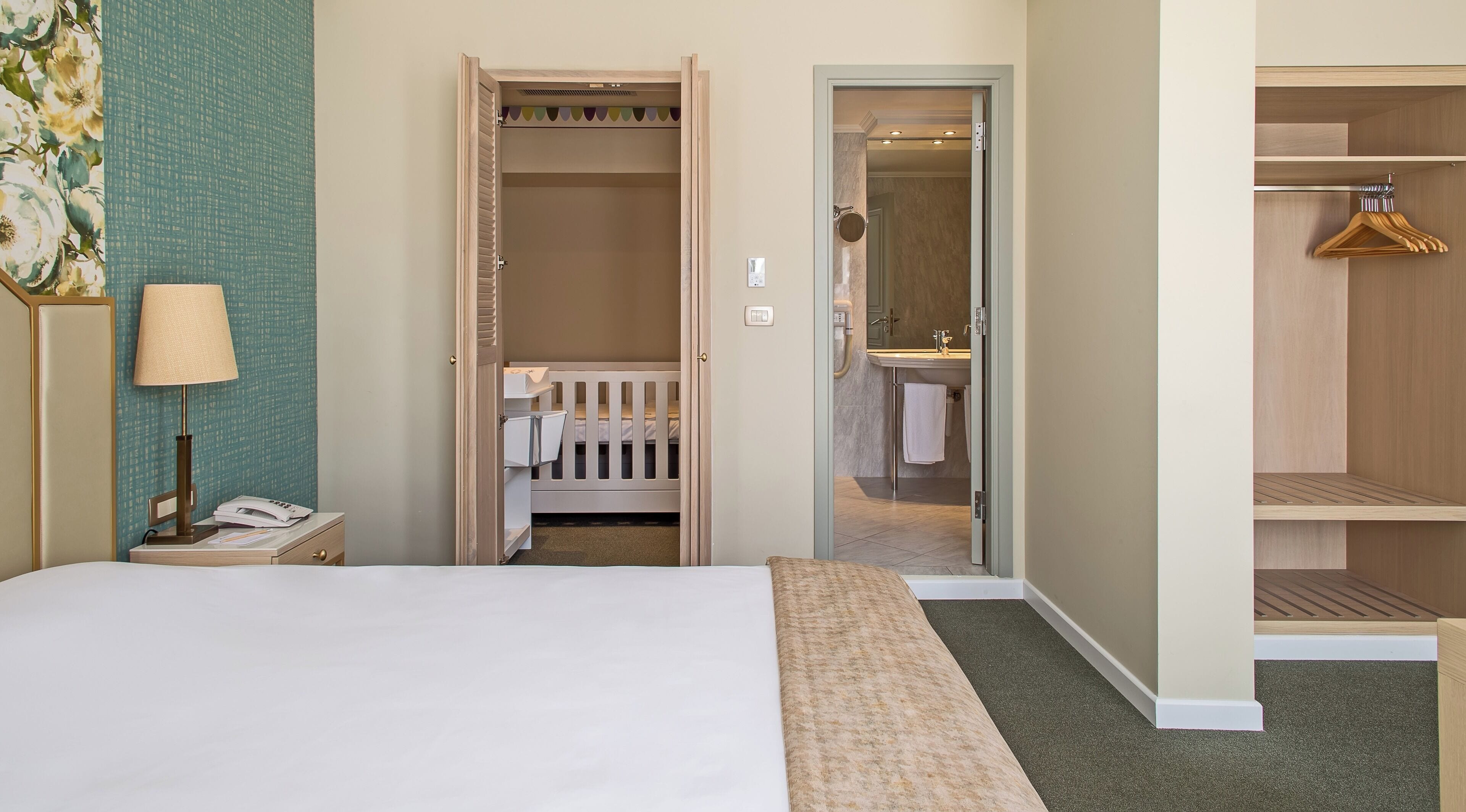 A hotel room with a bed, a bedside lamp, a crib, a closet, and a bathroom.