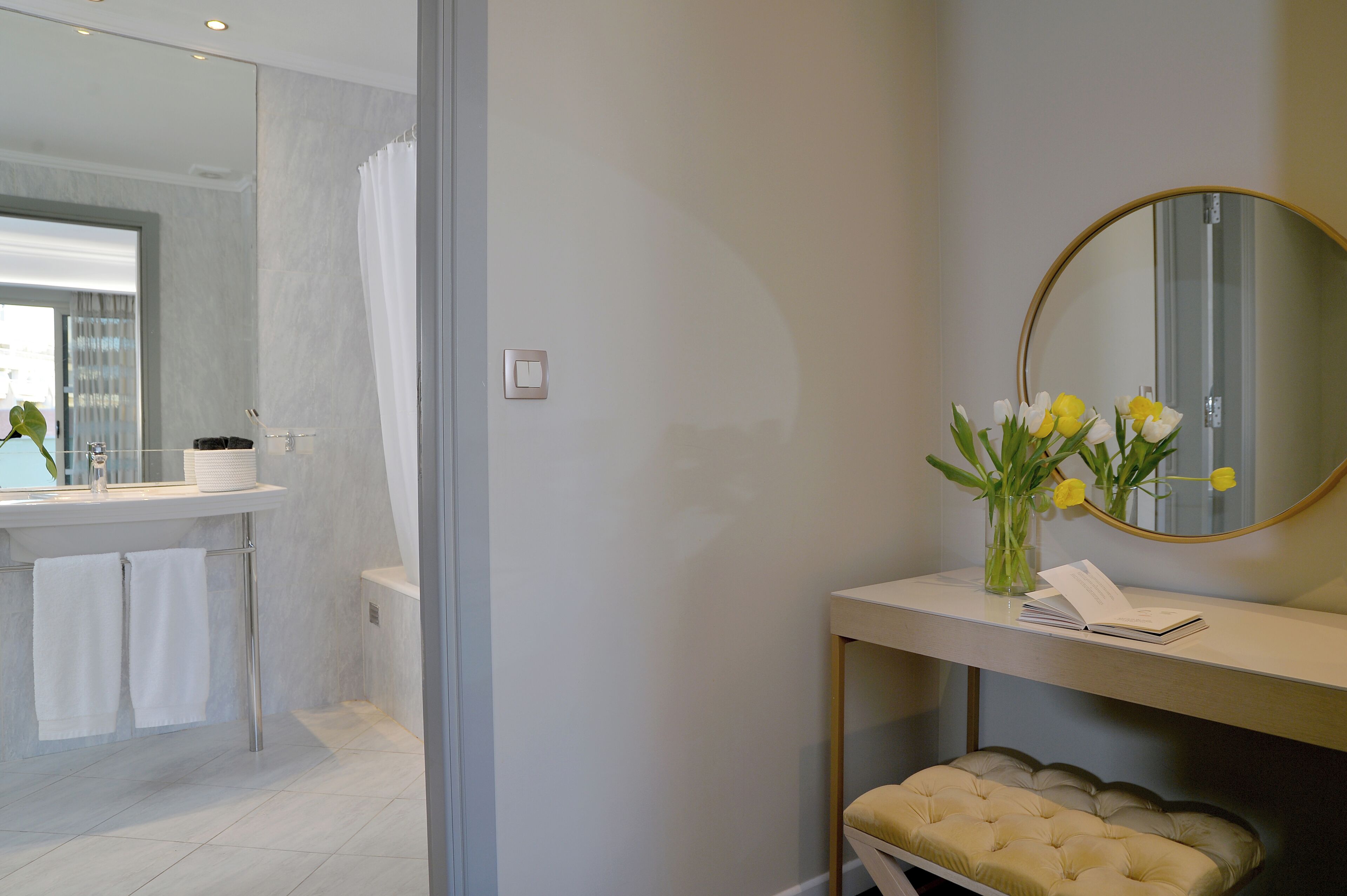 A bathroom with a mirror, a vanity with a vase of flowers, and a bench.