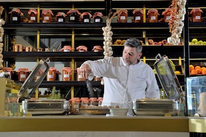 Uma pessoa vestindo uniforme branco em um balcão, com diversos alimentos e recipientes ao fundo.
