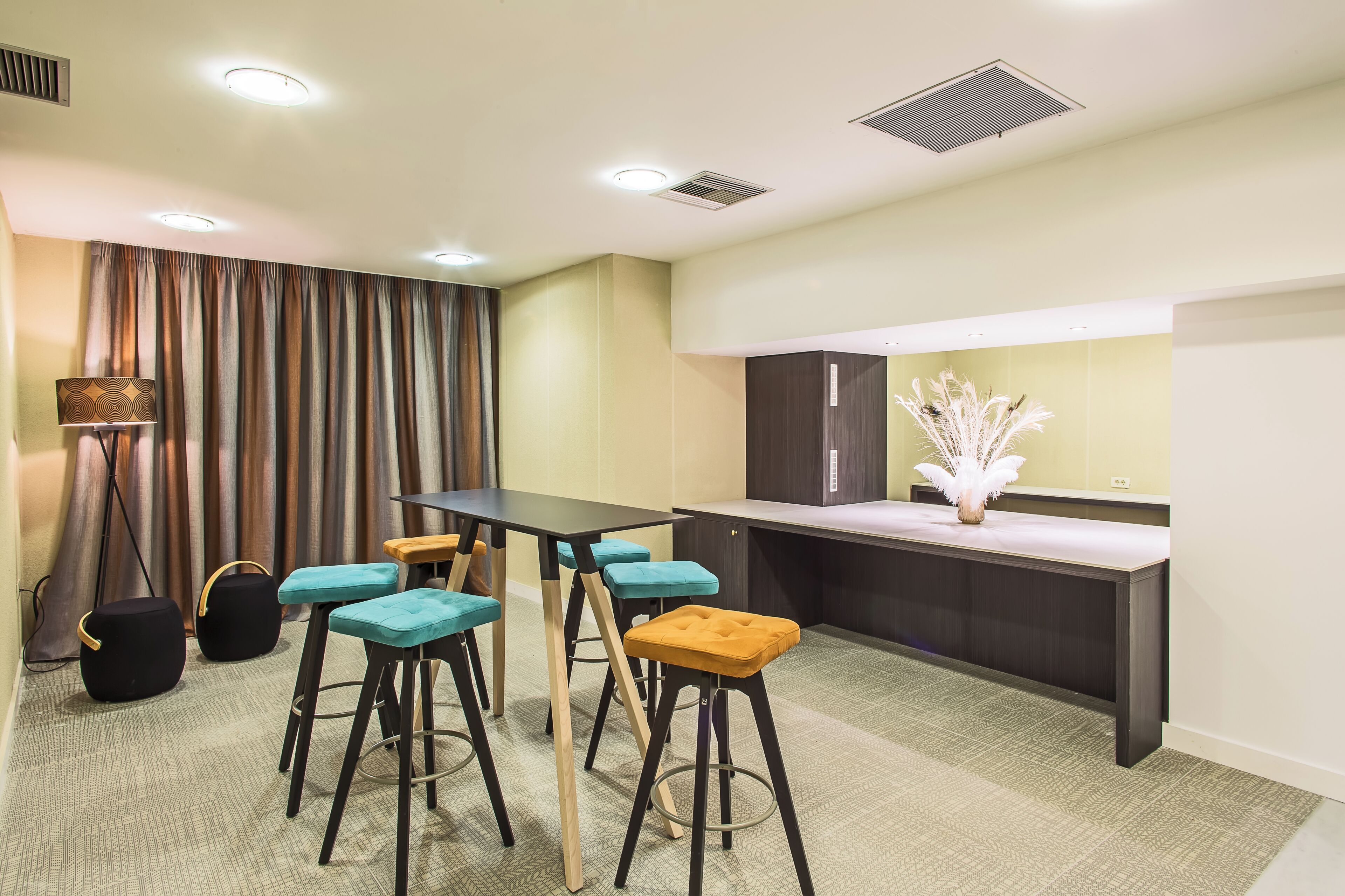 A conference room with a table, four stools, a bench, a vase with white feathers, and a lamp.