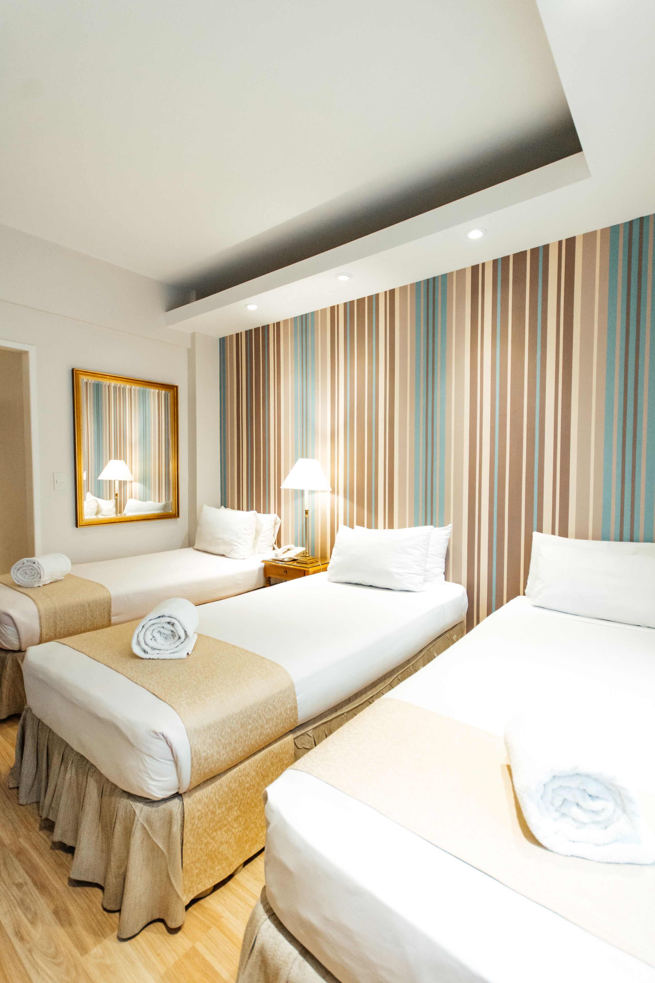 A hotel room with three single beds, a large mirror, striped wallpaper, and a wooden floor.
