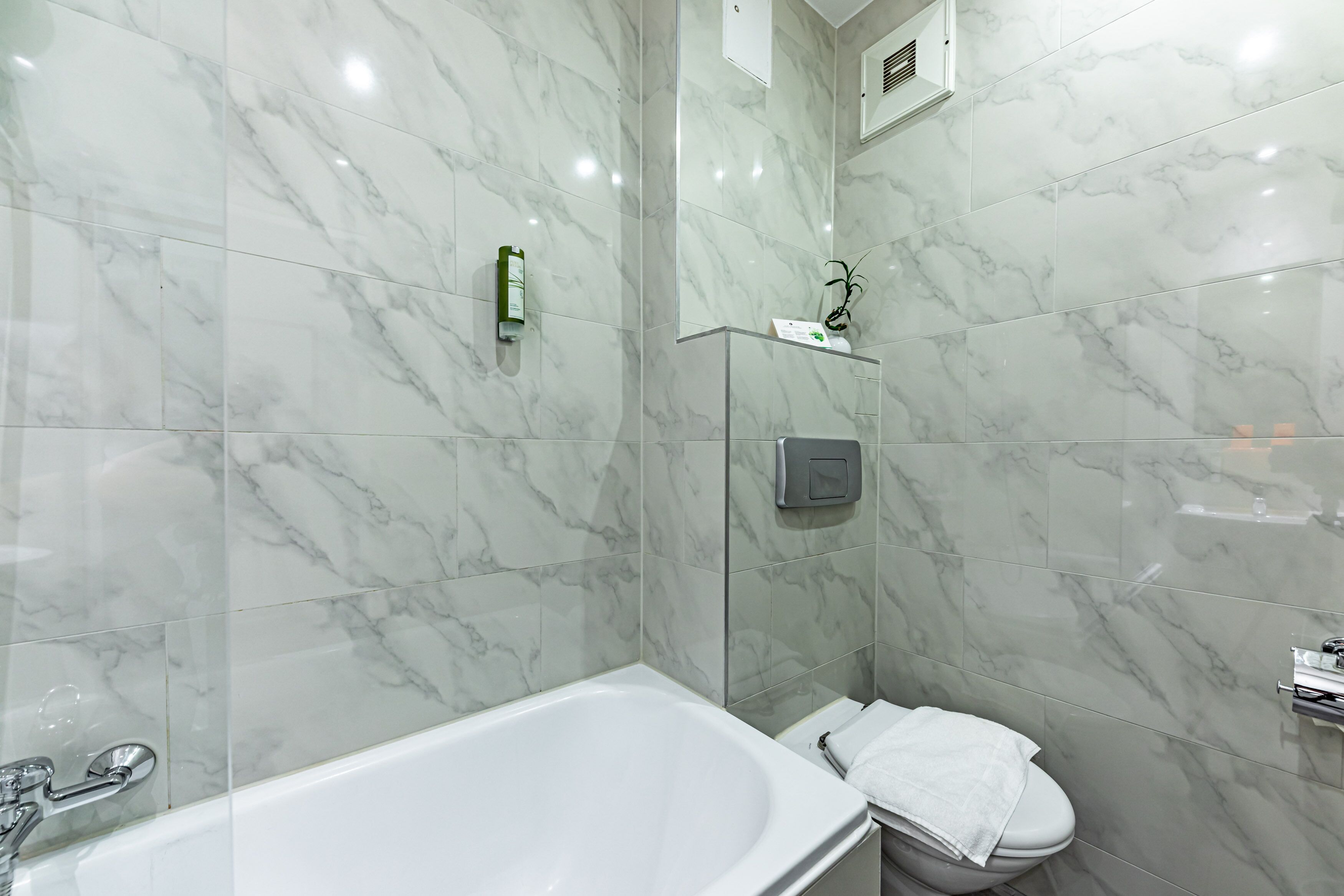 A bathroom with a marble-tiled wall, a white bathtub, a toilet, and a small shelf with toiletries.