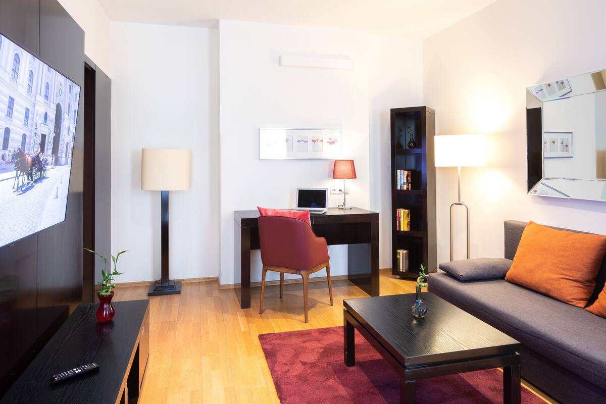 A modern living room with a flat-screen TV, a desk with a computer, a red chair, a grey sofa, and a red rug.