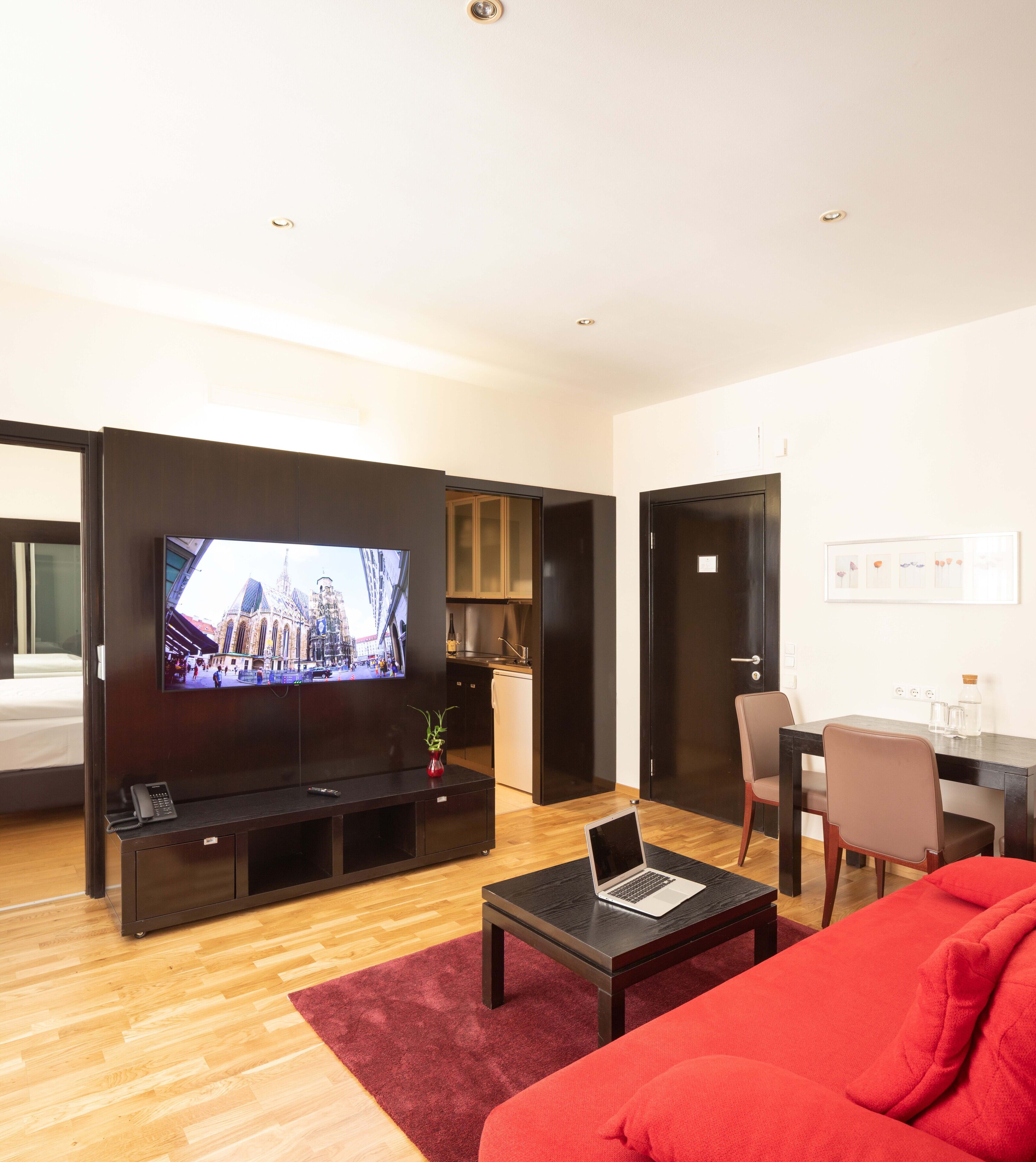 A modern living room with a flat-screen TV, a laptop on a coffee table, a red sofa, and a kitchen area in the background.