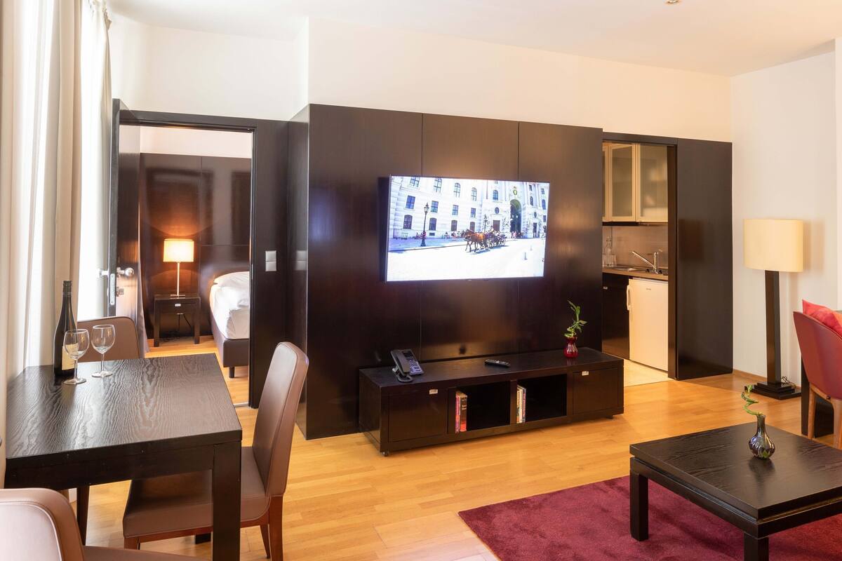 A hotel room with a TV, a dining table, chairs, a sofa, a small cabinet, a red rug, and a mirror.