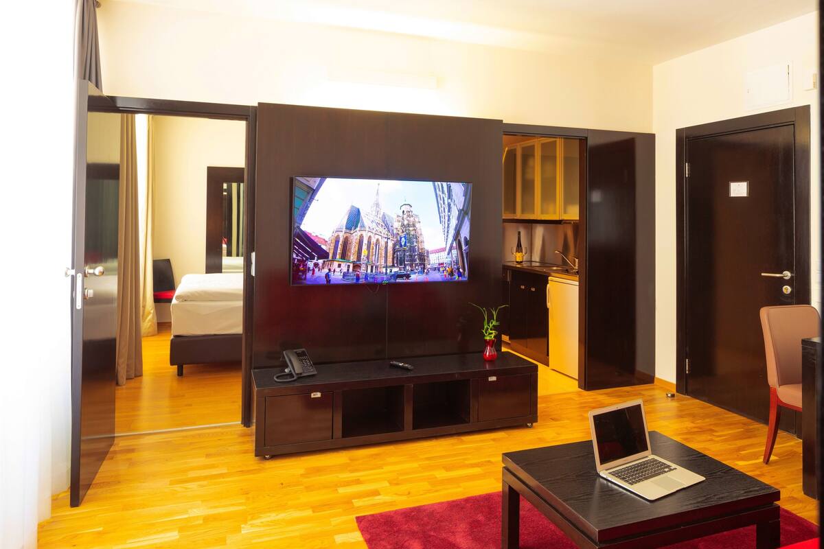 A modern hotel room with a large flat-screen TV, a bed, a red sofa, a wooden coffee table, and a laptop.