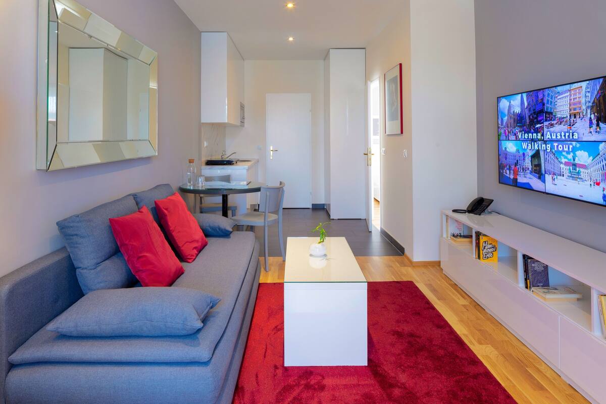 A modern living room with a grey sofa, red cushions, a white coffee table, and a red rug. A flat-screen TV on the wall displays a walking tour of Vienna, Austria.
