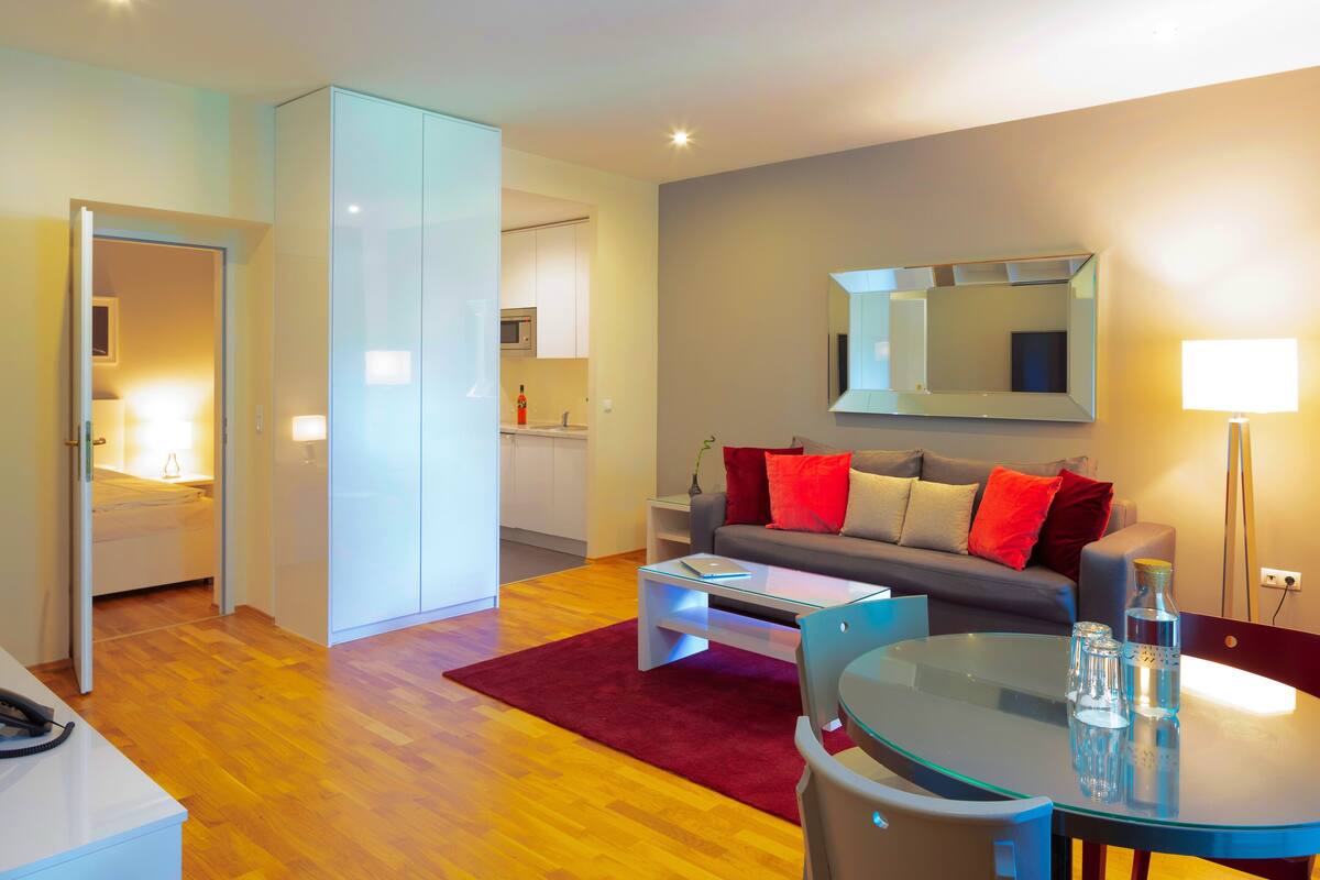 A modern living room with a glass coffee table, a sofa with red cushions, a dining area with a glass table, and a kitchen in the background.