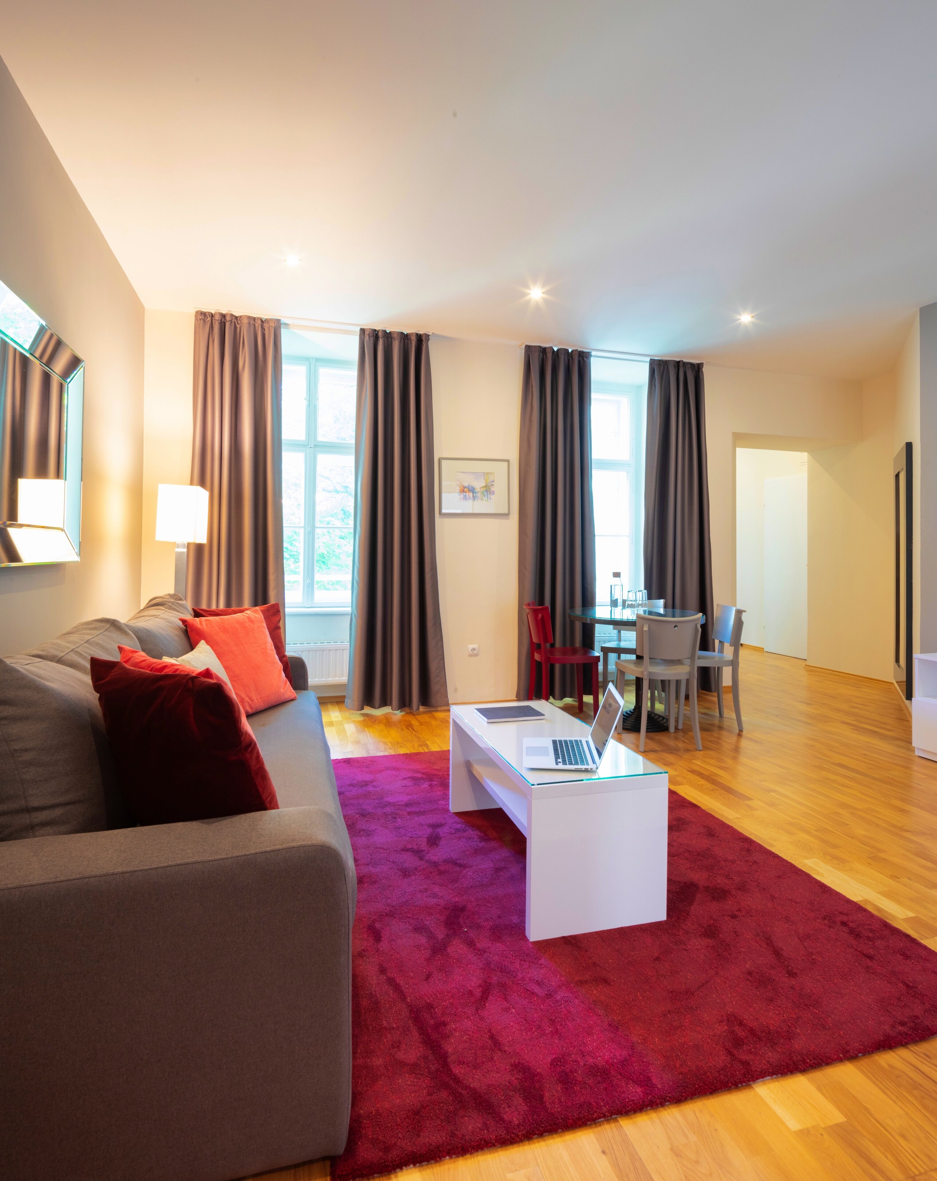 Ein modernes Wohnzimmer mit einem grauen Sofa, roten Kissen, einem weißen Couchtisch mit einem Laptop, einem roten Teppich und einem Essbereich mit Stühlen und einem Tisch.