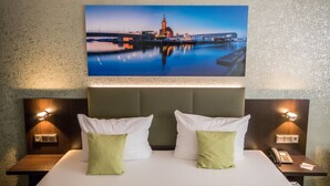 Habitación de hotel con una cama grande, dos almohadas, una mesita de noche con un teléfono y un cuadro de un paisaje urbano colgado en la pared.