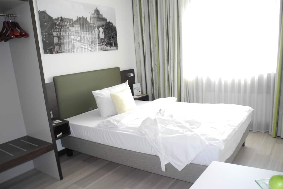 A hotel room with a neatly made bed, a nightstand with a lamp, a framed picture on the wall, and a coat rack with red shoes.