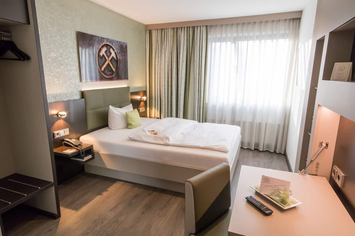 A hotel room with a bed, bedside table, nightstand, and a view of the outside through curtains.
