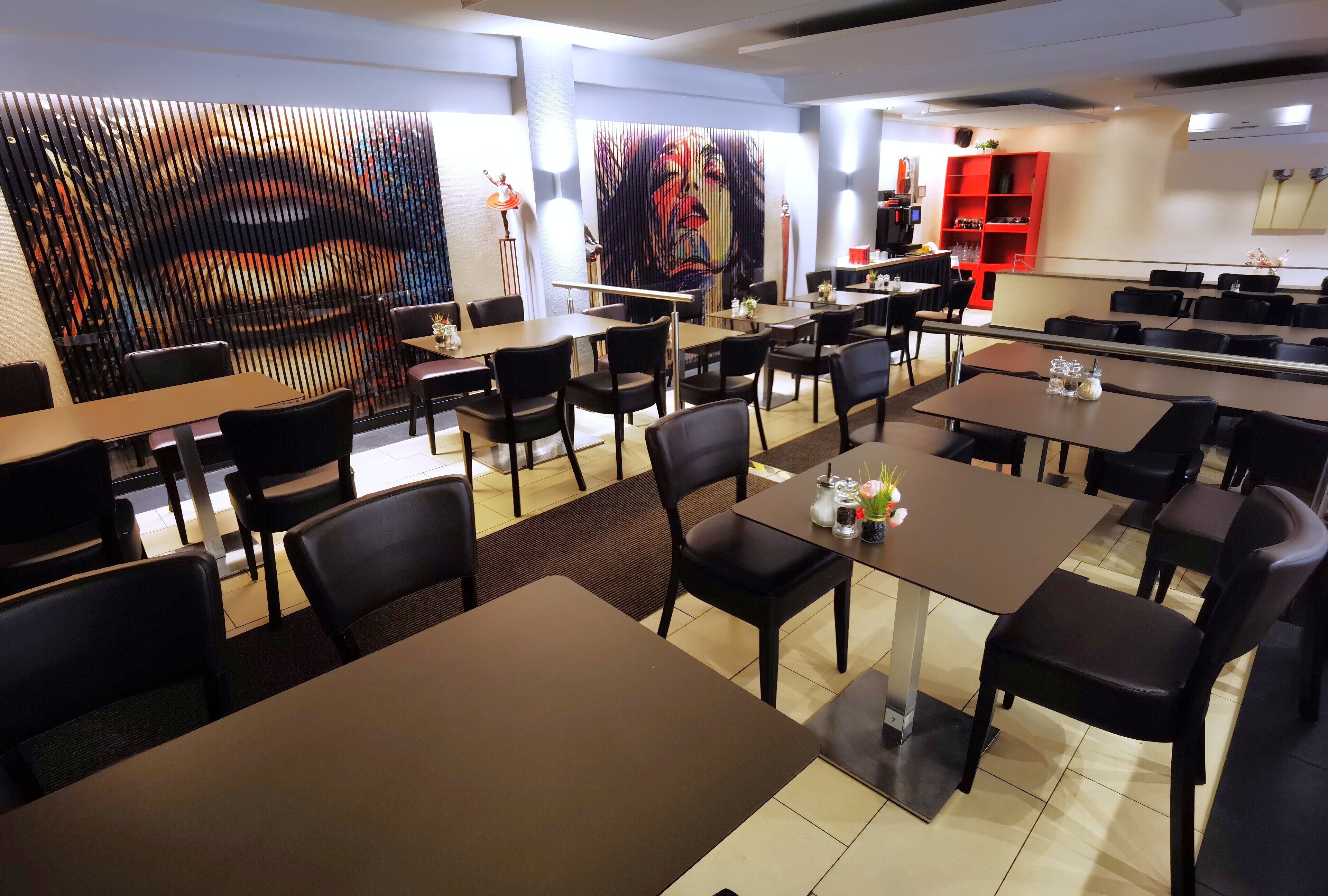 A restaurant with multiple tables and chairs, a large abstract art piece on the wall, and a red shelving unit in the background.