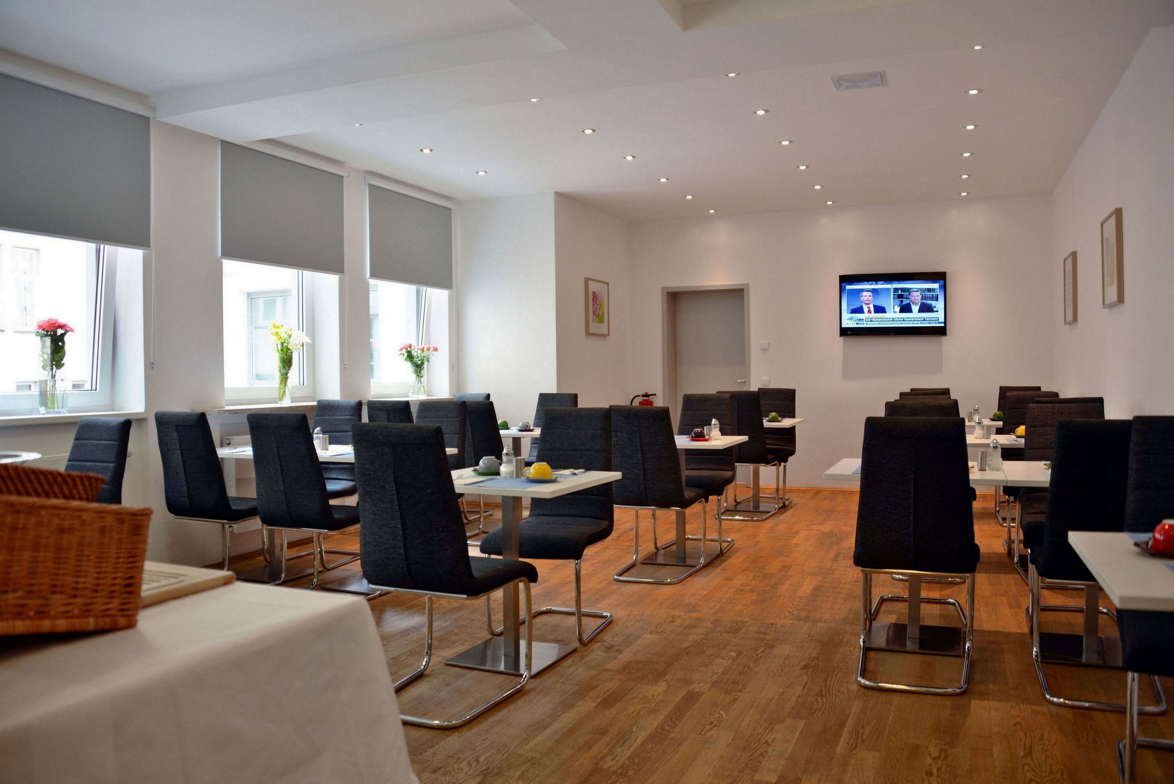 A modern dining area with multiple tables and chairs, large windows, and a television screen on the wall.