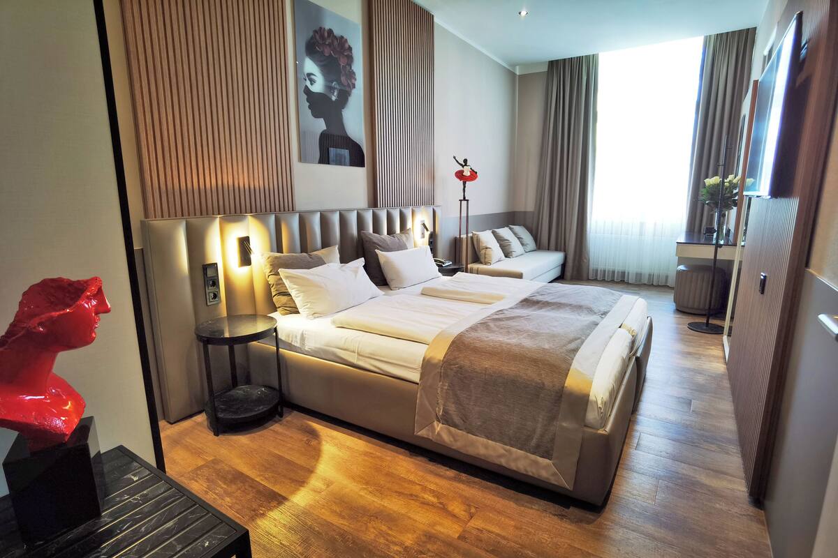A modern hotel room with a large bed, bedside tables, a red sculpture, and a large window with curtains.